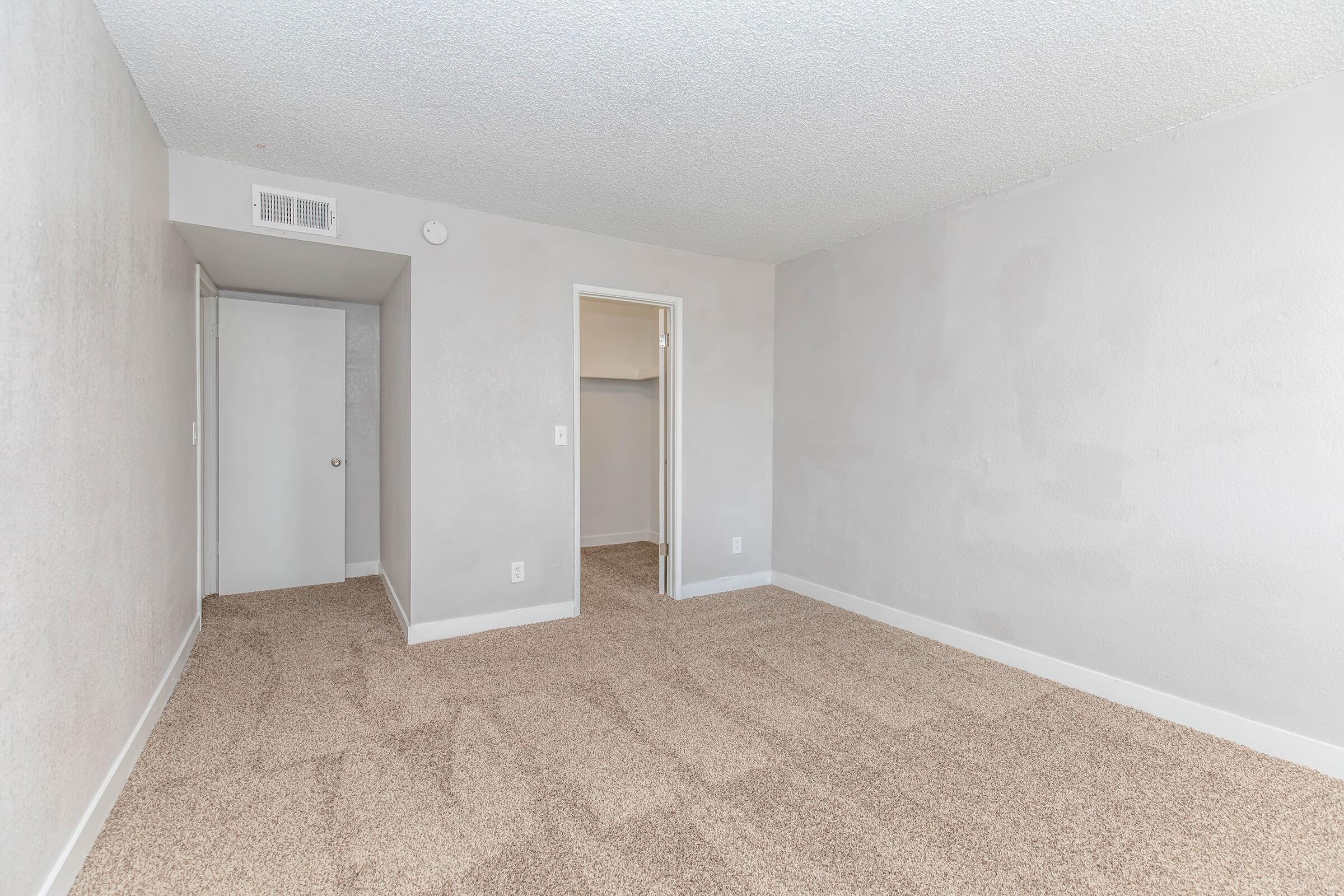 A spacious, empty room with light gray walls and carpeted flooring. There is a closet on the left and a doorway leading to another small room. The space is well-lit with natural light, creating a bright and inviting atmosphere.