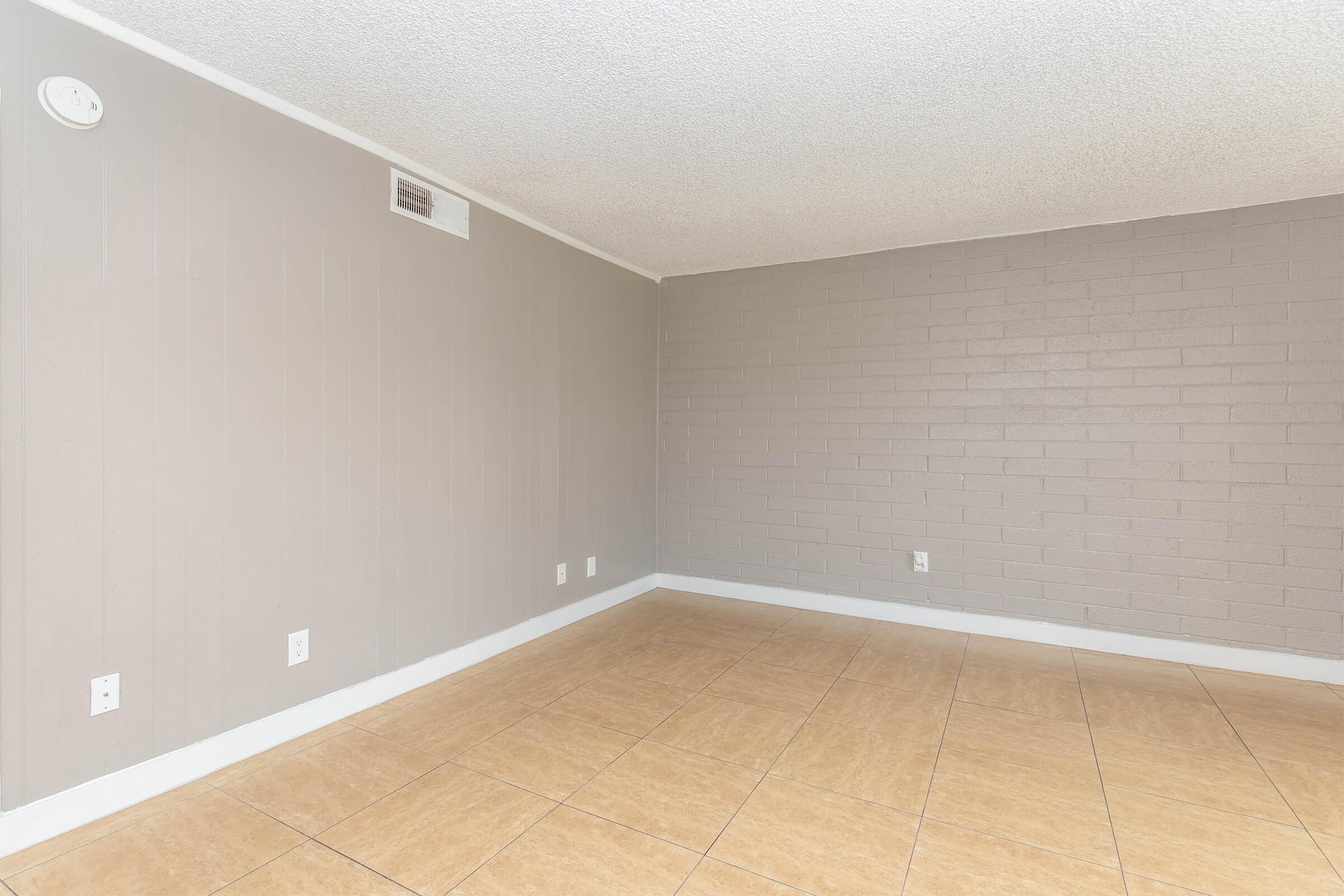 Empty room with beige tiled flooring, featuring a light gray textured wall on one side and a smooth light gray wall on the other. There are electrical outlets on the walls, and natural light illuminates the space through the ceiling. The overall atmosphere is minimalistic and clean.