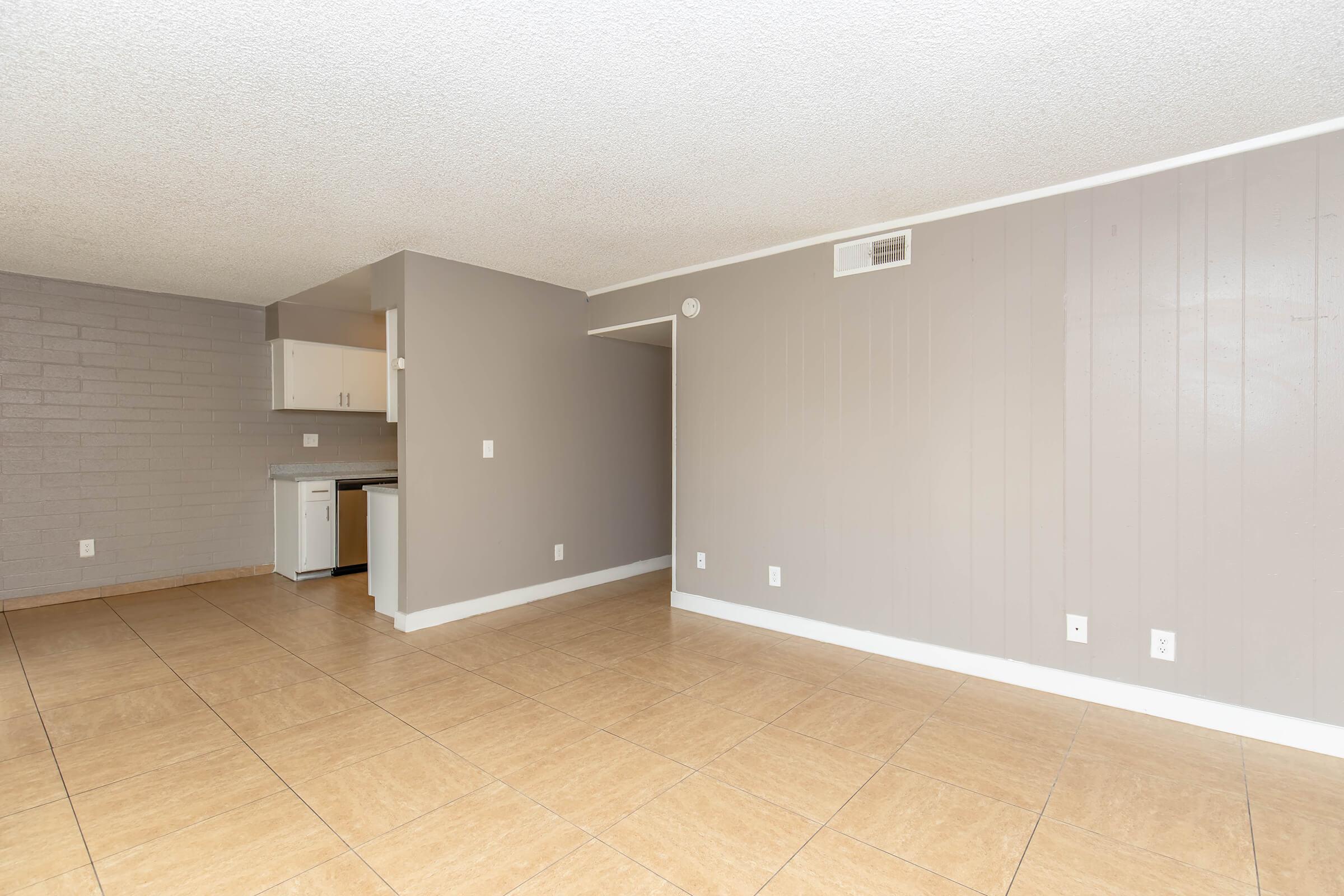 A spacious, light-filled room featuring neutral-colored walls and tiled flooring. The open layout connects to a small kitchen area with white cabinets in the background, adding to the modern and clean aesthetic. Natural light highlights the simplicity and versatility of the space.