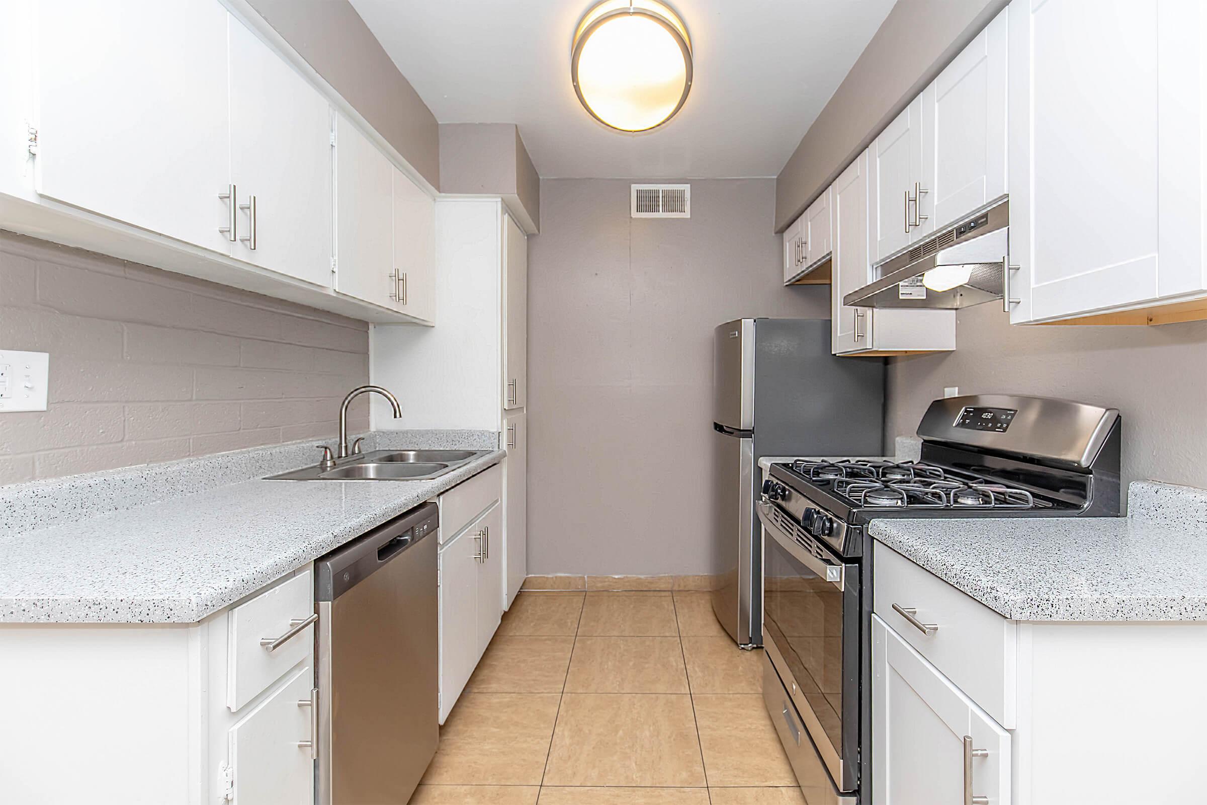 A modern kitchen featuring white cabinetry, a stainless steel gas stove, a silver refrigerator, and a dishwasher. The countertops are light gray with a speckled design, and the walls are painted in a neutral tone. The floor is tiled with beige tiles, and there is a ceiling light fixture above.