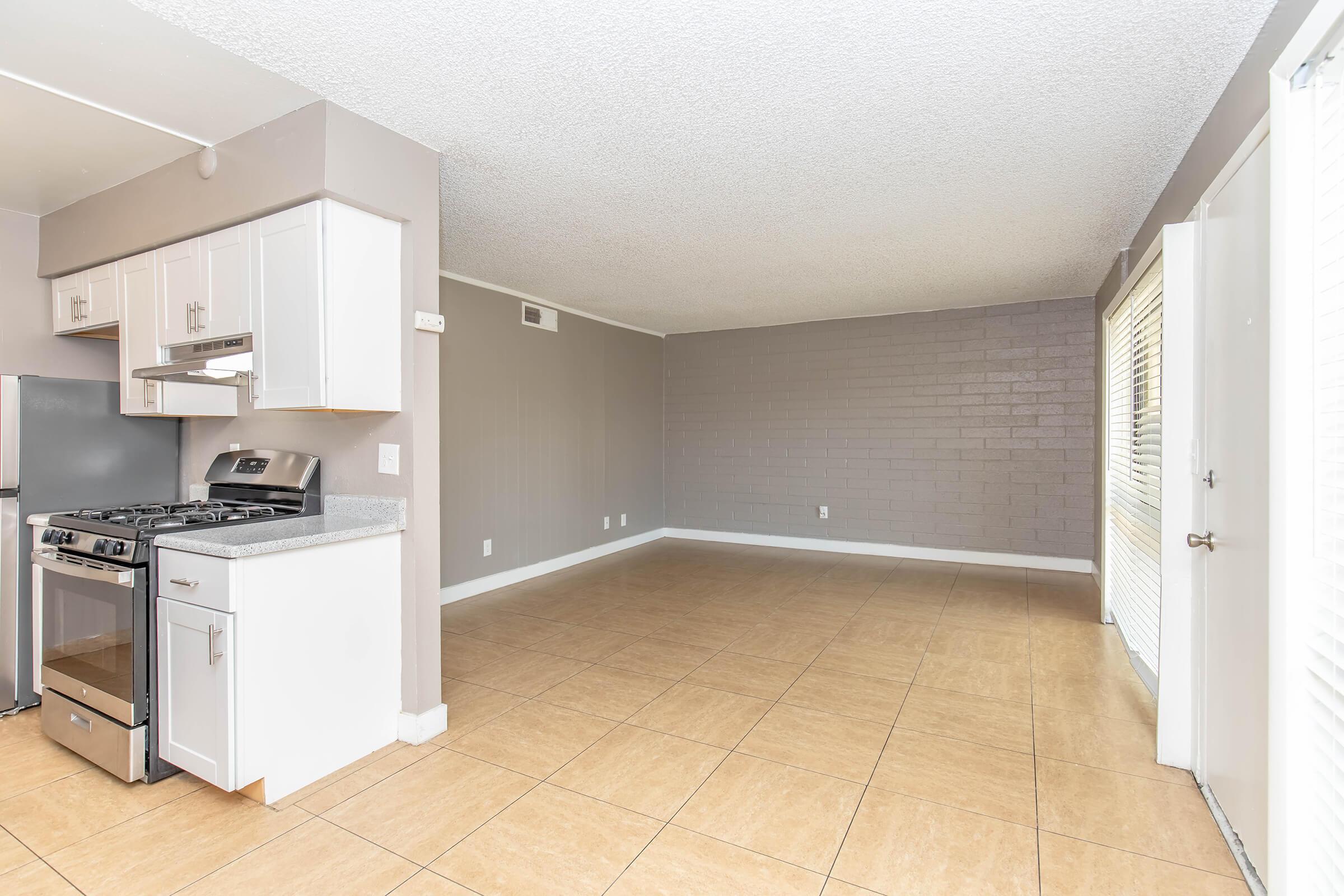 A spacious, empty living room with beige tiled flooring and gray walls. On one side, there is a modern kitchen with white cabinets and stainless steel appliances, including a gas stove. Large windows let in natural light, and there is a door that opens to the outside. The room has a clean, minimalist feel.