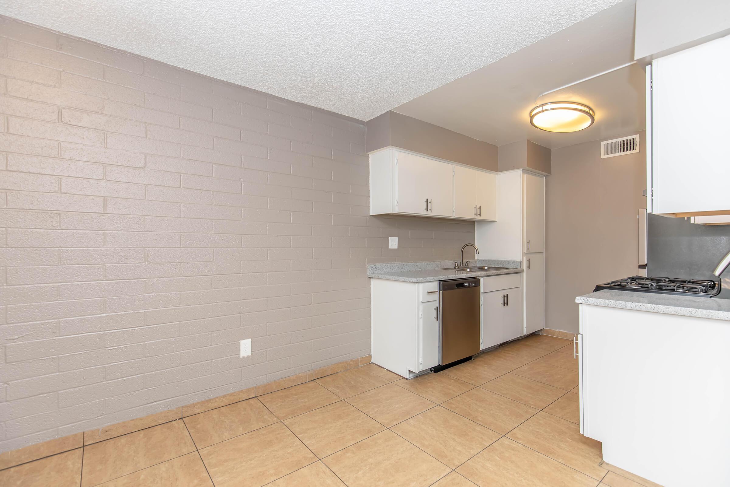 A spacious kitchen featuring light-colored cabinetry, a stainless steel dishwasher, and a double-basin sink. The walls have a textured gray finish, and the floor is tiled in a light brown hue. A ceiling light fixture provides overhead illumination, enhancing the open feel of the area.