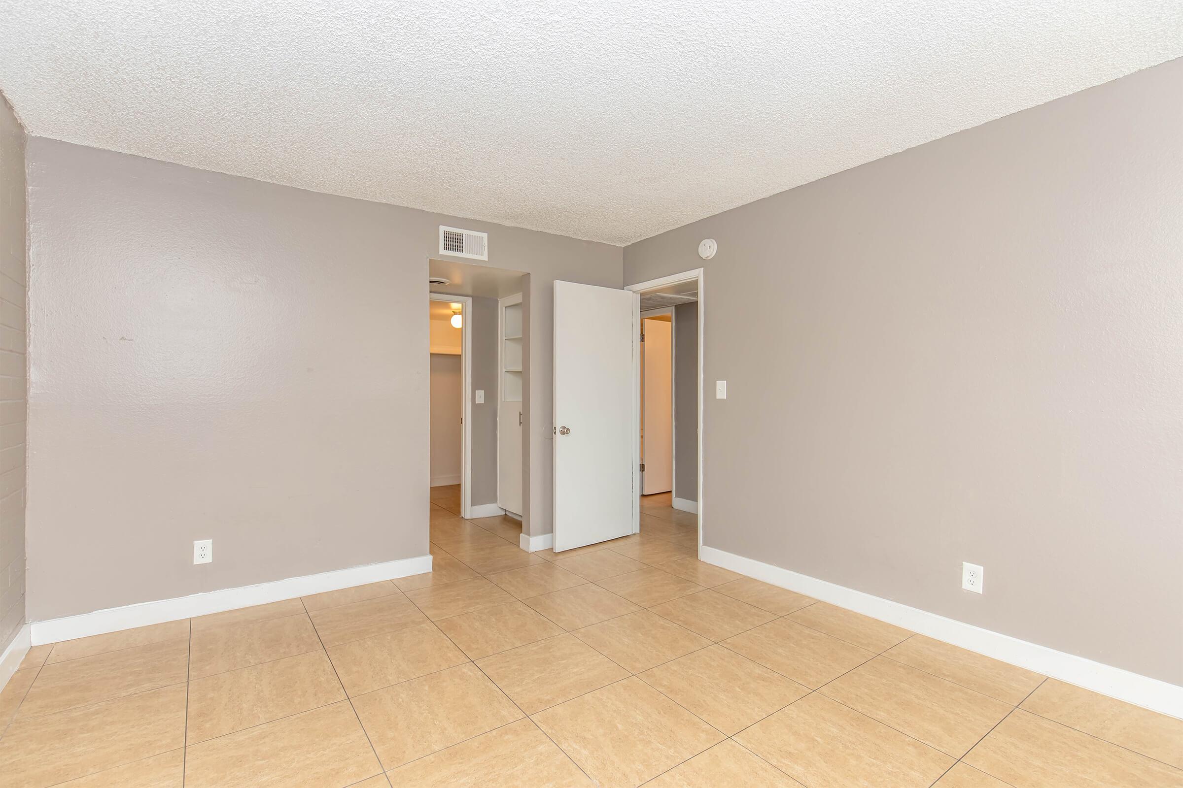 An empty room with light gray walls and tile flooring. There is a doorway leading to a closet and another room. The ceiling has a textured finish, and there is natural light coming from an unseen window, creating a bright and open atmosphere.