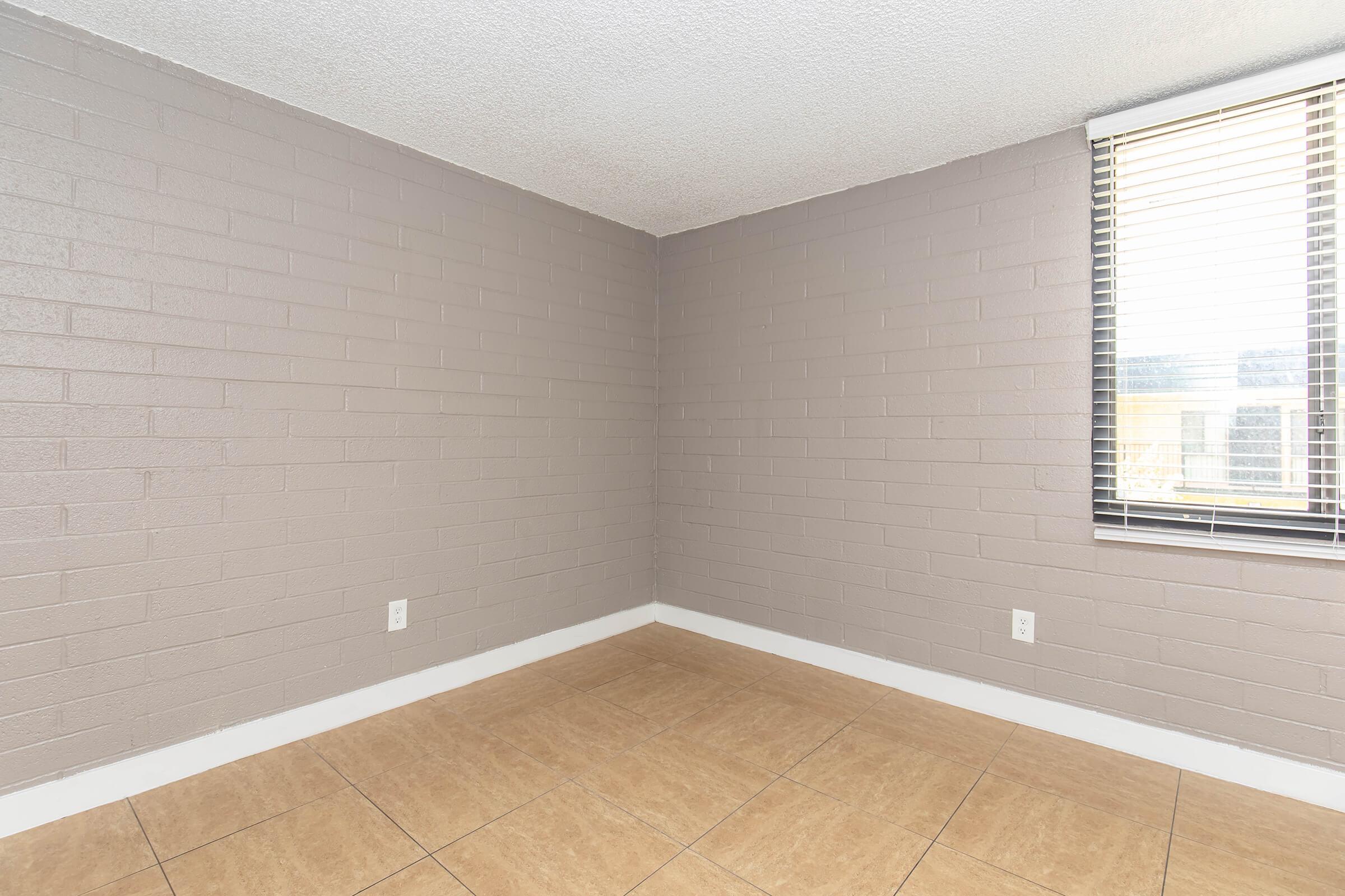 Empty room with light gray brick walls and a window featuring horizontal blinds. The floor is tiled in a light brown pattern. There are no furnishings or decorations, creating a spacious and minimalist appearance.