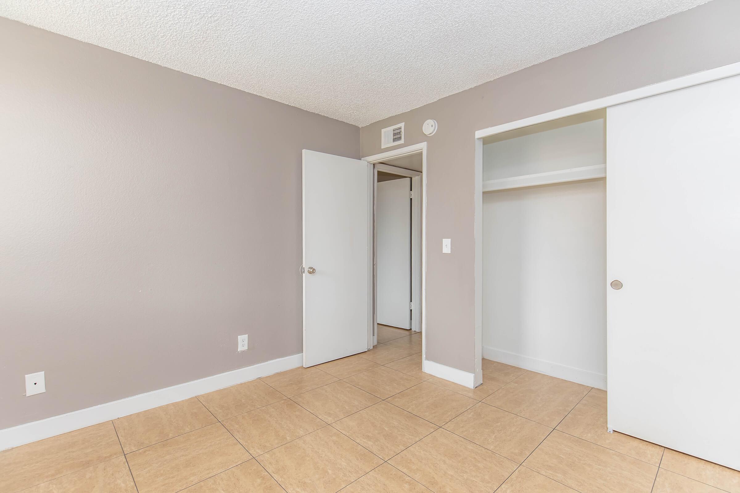 Empty room with light gray walls and tiled floor. A closed door on the left leads to another area, while two closet doors are visible on the right. The room is well-lit, showcasing a simple and clean aesthetic with minimal furnishings.