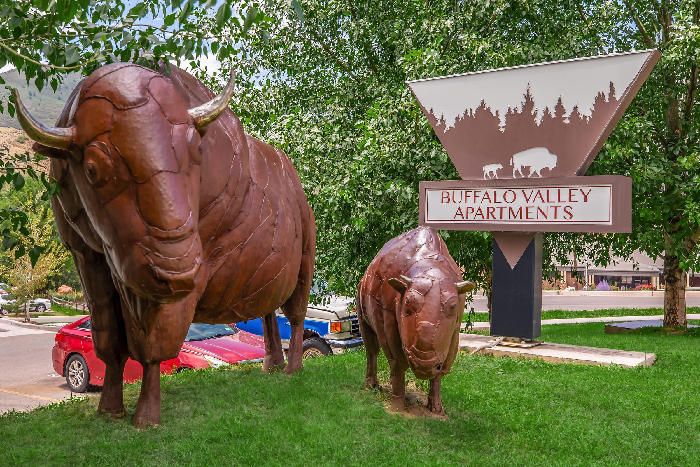 Two large bronze bison sculptures stand in front of a sign for Buffalo Valley Apartments, framed by lush green trees. A parked truck and an open parking area are visible in the background, highlighting a scenic, welcoming environment. The sign features a silhouette of bison and forest.