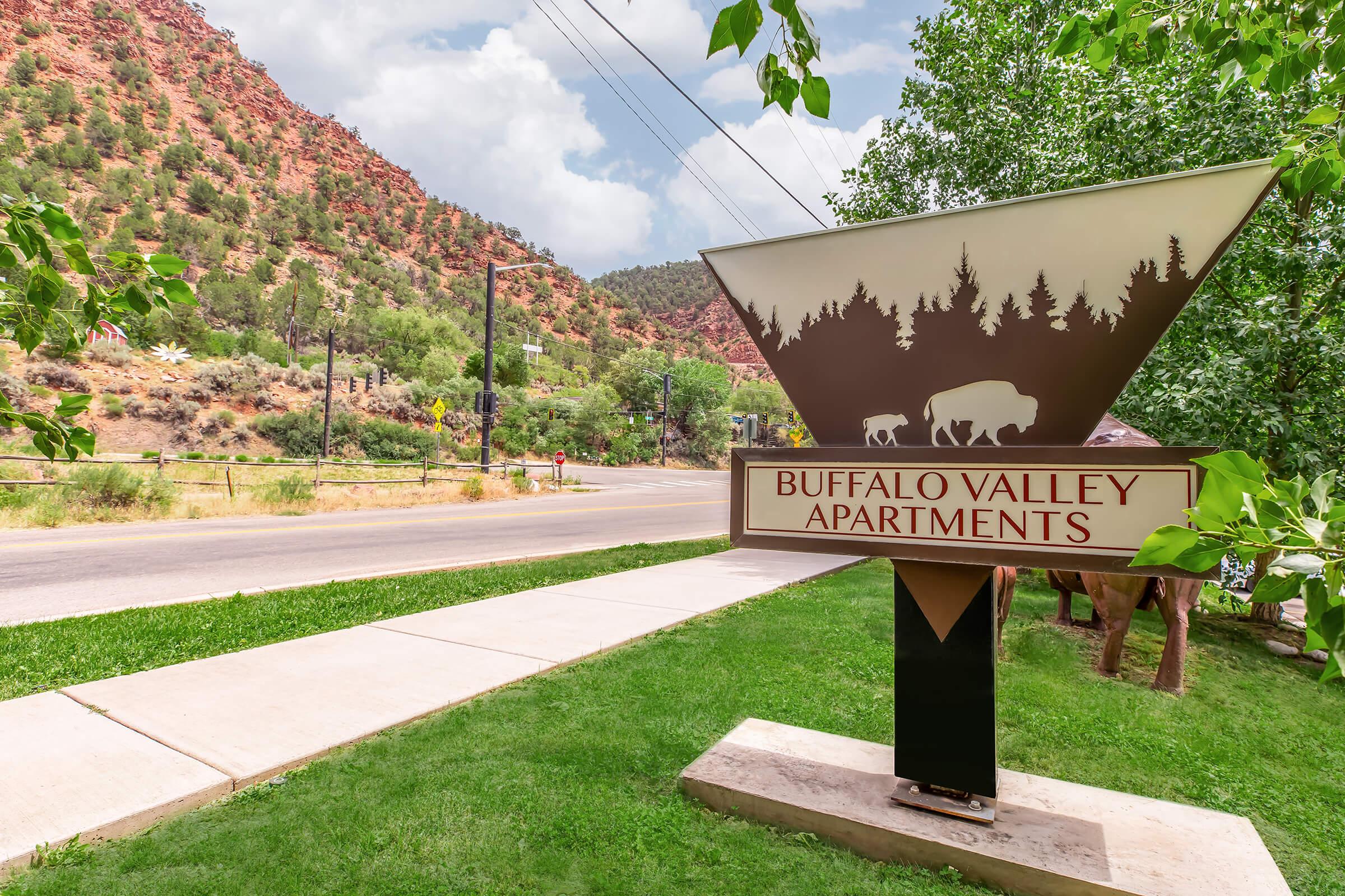 Sign for Buffalo Valley Apartments with a scenic view of green trees and rocky, mountain terrain in the background. The sign features a silhouette of bison and forest elements, standing on a landscaped area next to a road. The sky is partly cloudy, adding to the natural ambiance.