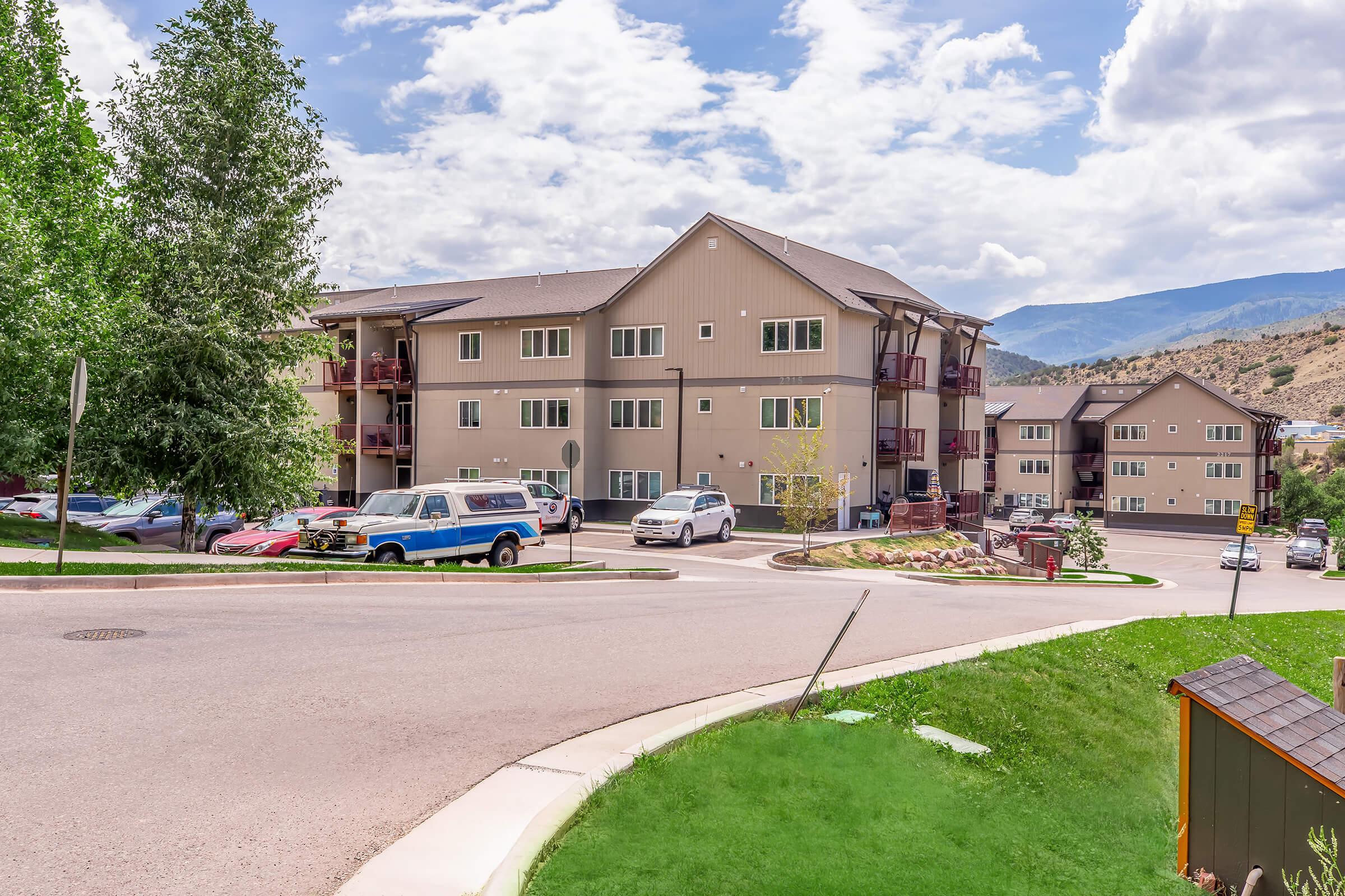 A view of a multi-story residential building situated on a street corner, surrounded by greenery and parked vehicles. In the background, hills and clouds are visible, indicating a scenic area. The building features balconies and a mix of modern architectural elements.