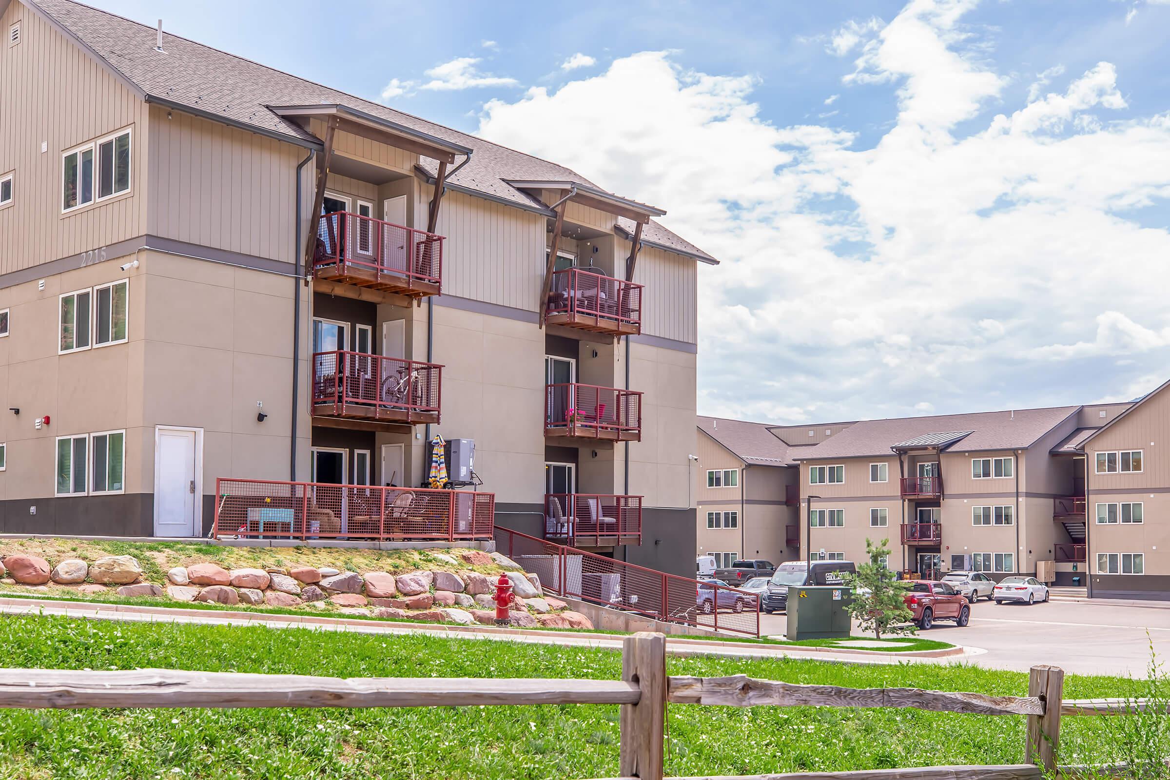 A modern apartment building with multiple balconies, set against a blue sky with scattered clouds. In the foreground, there is a grassy area with a wooden fence and a fire hydrant. The background shows additional apartment units and parking spaces.