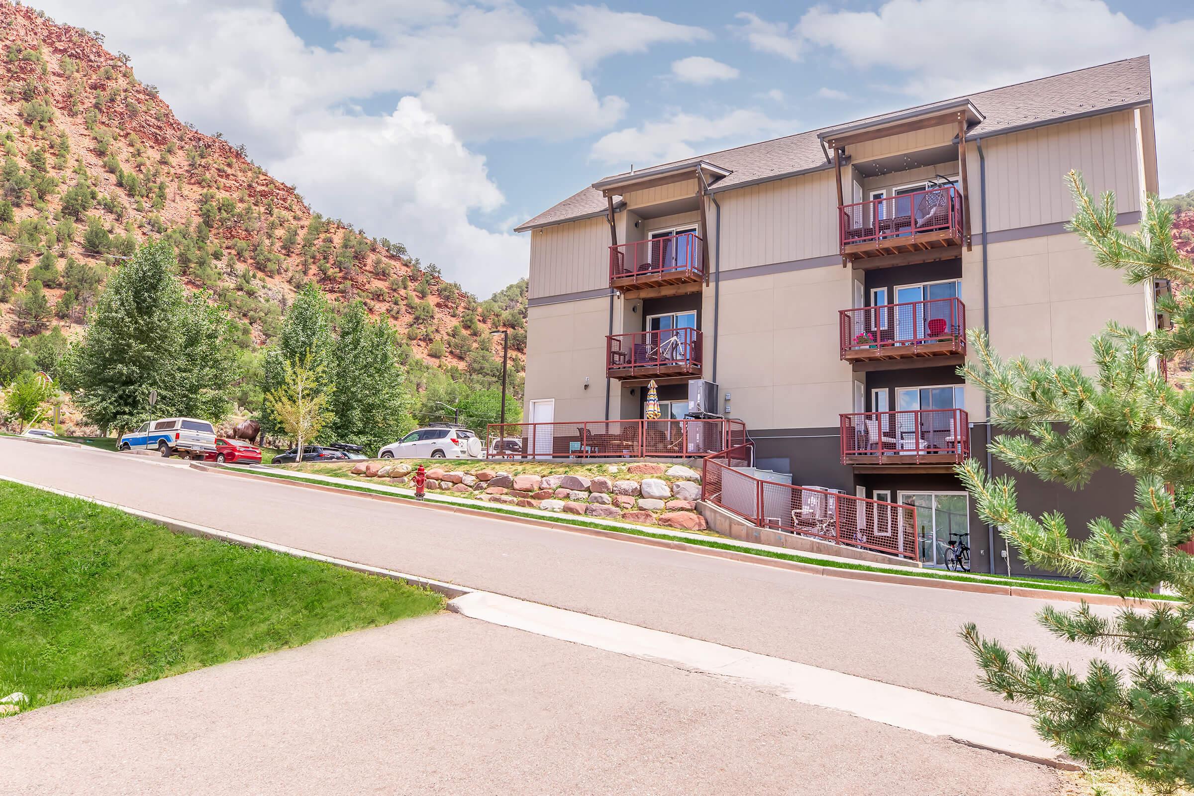 A modern multi-story building situated on a sloped street, surrounded by lush greenery and rocky hills. The structure features balconies on the upper floors and is backed by a mountainous landscape, with a few vehicles parked nearby and a clear sky above.