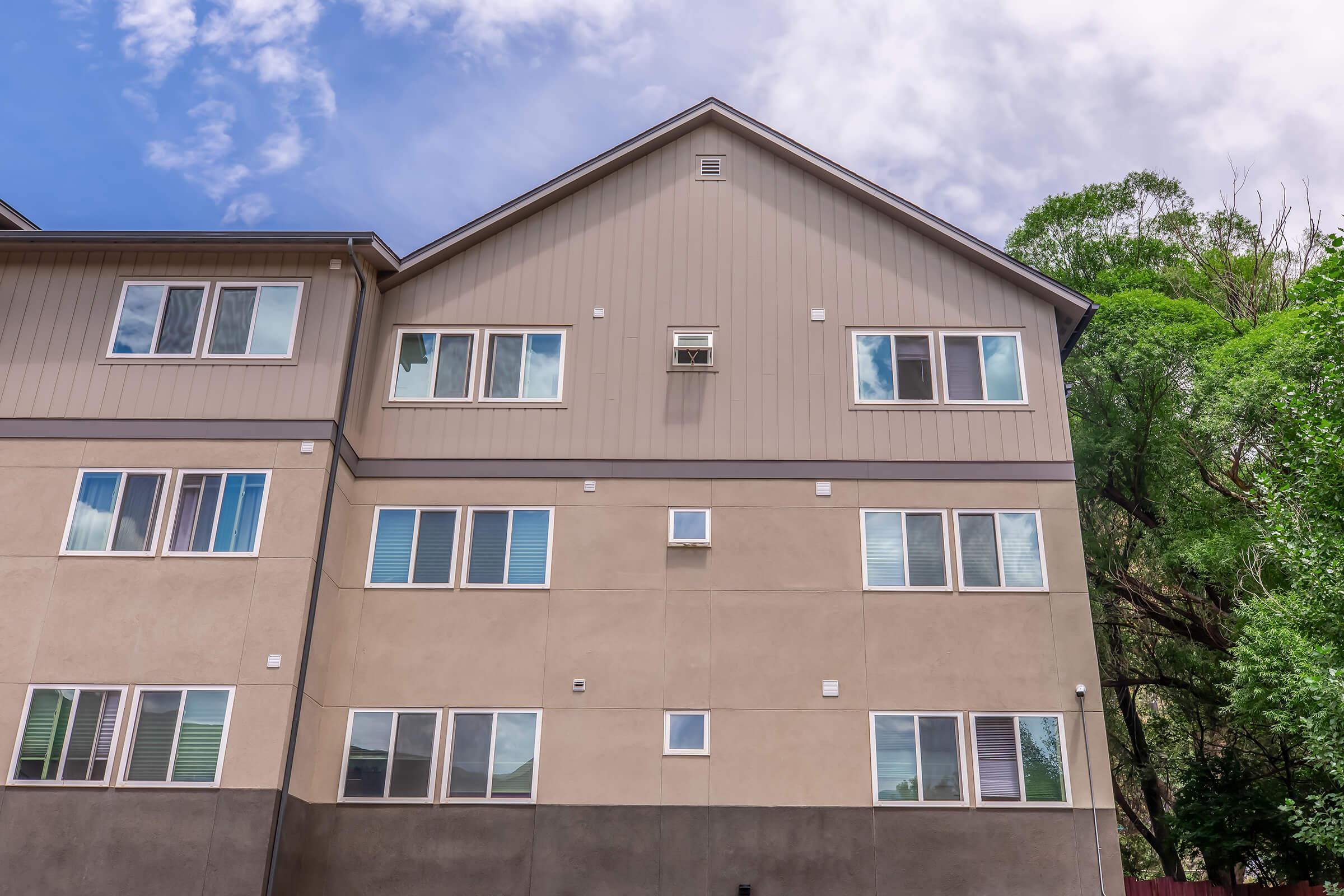 A two-story building with a light-colored exterior and multiple windows. The upper level features several rectangular windows under a sloping roof, while the lower level has a mix of larger and smaller windows. Green trees are visible in the background under a blue sky with some clouds.