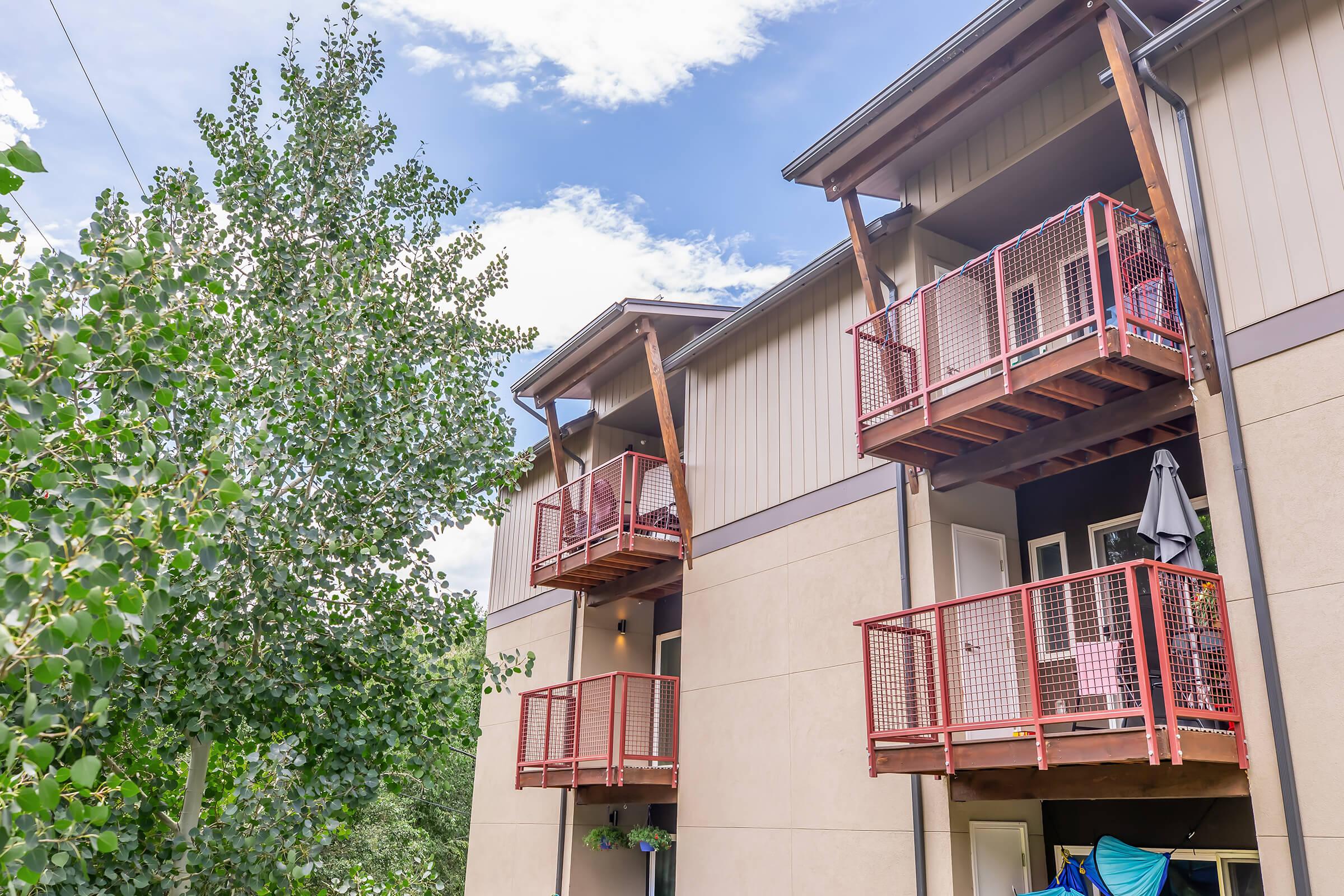 Exterior view of a three-story building featuring balcony spaces with red metal railings, surrounded by greenery. A large tree is visible on the left, and some laundry items are hanging below the balconies, indicating a residential setting. The sky is partly cloudy with blue and white hues.
