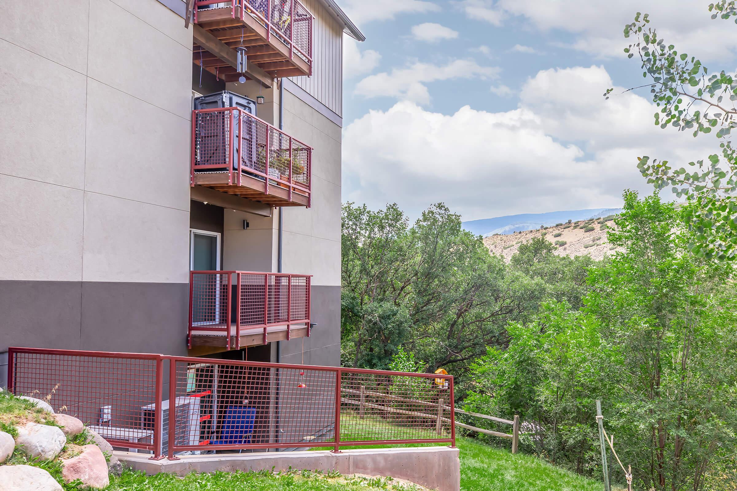A multi-story building with balconies surrounded by greenery. The building features a mix of light and dark exterior colors. In the foreground, there is a grassy area with rocks and a fenced section. The sky is partly cloudy, and hills are visible in the background.