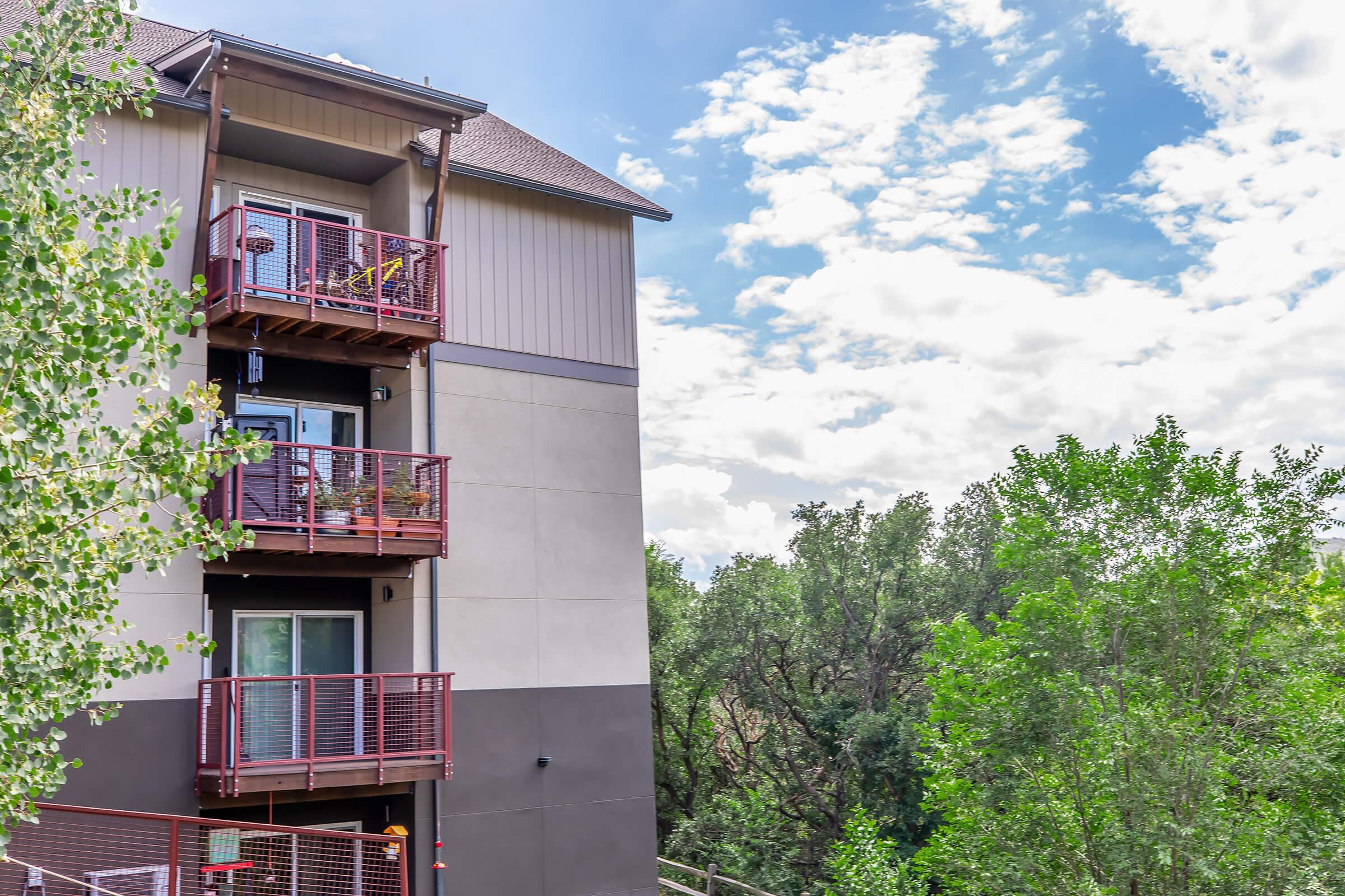 A multi-story building featuring multiple balconies with red railings. The design includes a mix of light and dark-colored exterior panels. In the background, lush greenery and a partly cloudy blue sky can be seen, contributing to a scenic view.