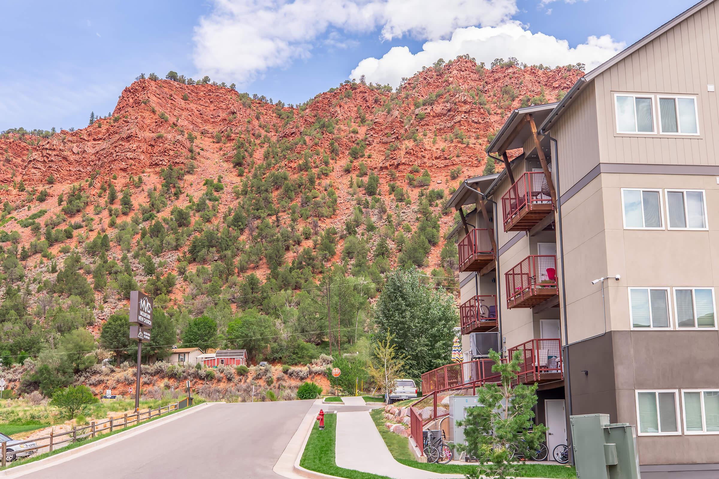 A modern multi-story building with balconies sits at the foreground, adjacent to a paved road. Behind it, vibrant red rocky hills rise under a blue sky with scattered clouds, creating a striking natural backdrop. Lush greenery and a few trees add to the scenic landscape.