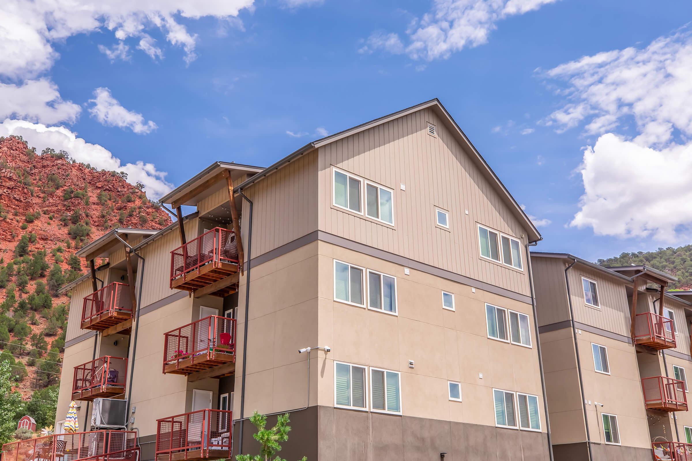 Three-story apartment building with beige and brown exterior, featuring several balconies with red railings. The building is set against a backdrop of green hills and a blue sky with white clouds.