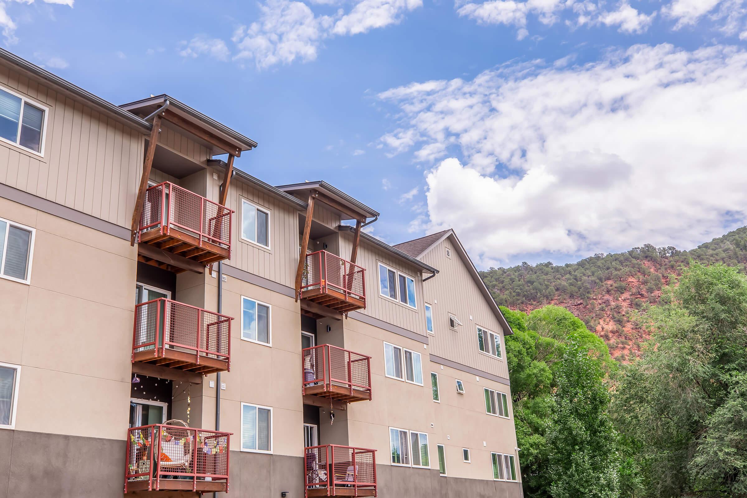 A multi-story apartment building with balconies, set against a backdrop of red rock formations and blue sky. Green trees are visible in the foreground, adding to the natural scenery. The image captures a modern residential area with a clear, sunny atmosphere.