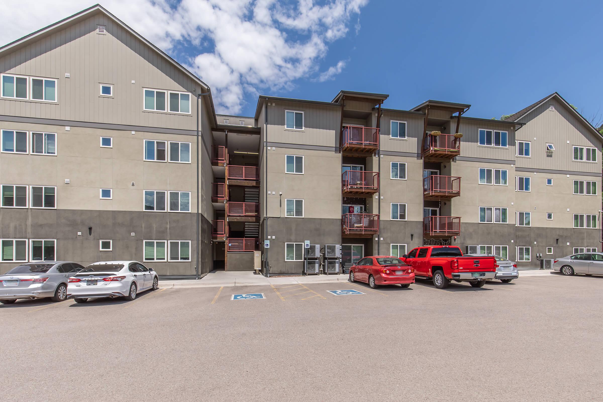 A modern multi-story residential building with balconies, featuring a mix of grey and beige siding. The foreground shows a parking lot with several vehicles, including a red truck and cars parked in designated spaces. The sky is mostly clear with some clouds.