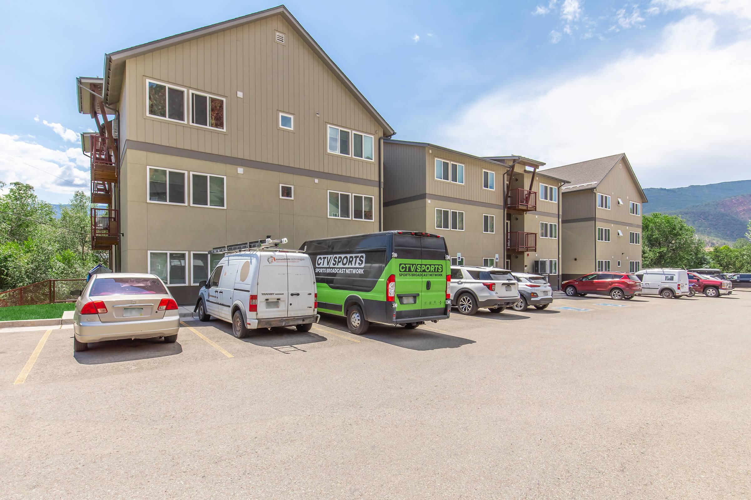 A modern multi-story apartment building with brown and beige siding, featuring balconies. Several parked vehicles, including a van with a green and black design labeled "CITY SPORTS," are visible in the parking lot. The backdrop includes trees and a blue sky.