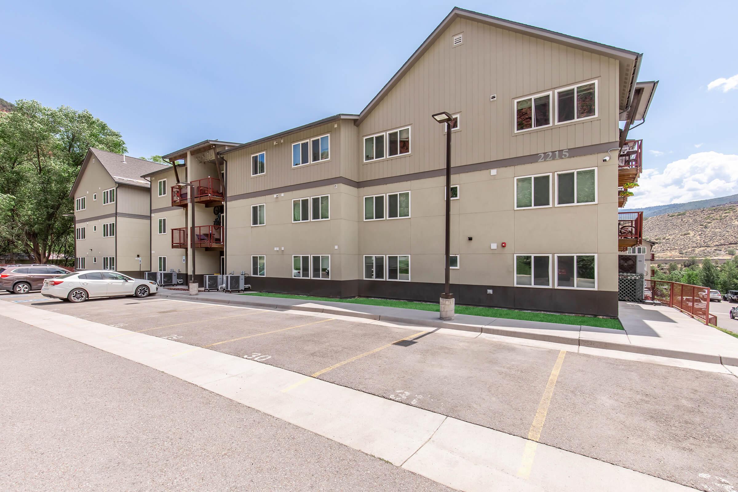 A modern multi-story apartment building with large windows and balconies, set against a clear blue sky. The building features light-colored siding and a well-maintained parking area with designated parking spaces. Lush trees are in the background, adding greenery to the environment.