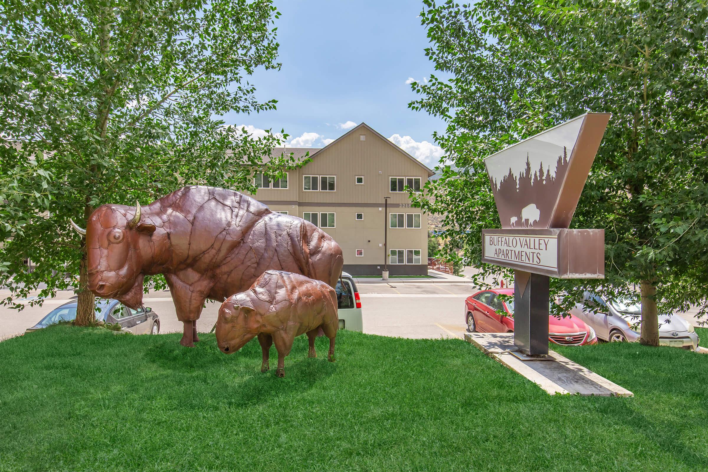 Two large buffalo sculptures, one adult and one calf, stand on green grass in front of the Buffalo Valley Apartments sign. The backdrop features a multi-story building surrounded by trees under a clear blue sky. The sign has a stylized image of a forest and the apartment name prominently displayed.