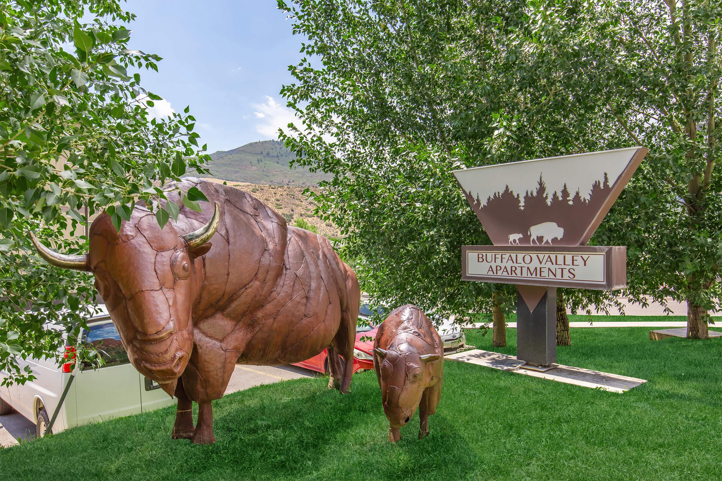 A large and a small bison sculpture made of metal are situated on a grassy area near a sign for Buffalo Valley Apartments. The sign features a triangular design with trees and a bison silhouette, surrounded by green foliage and mountains in the background under a clear sky.