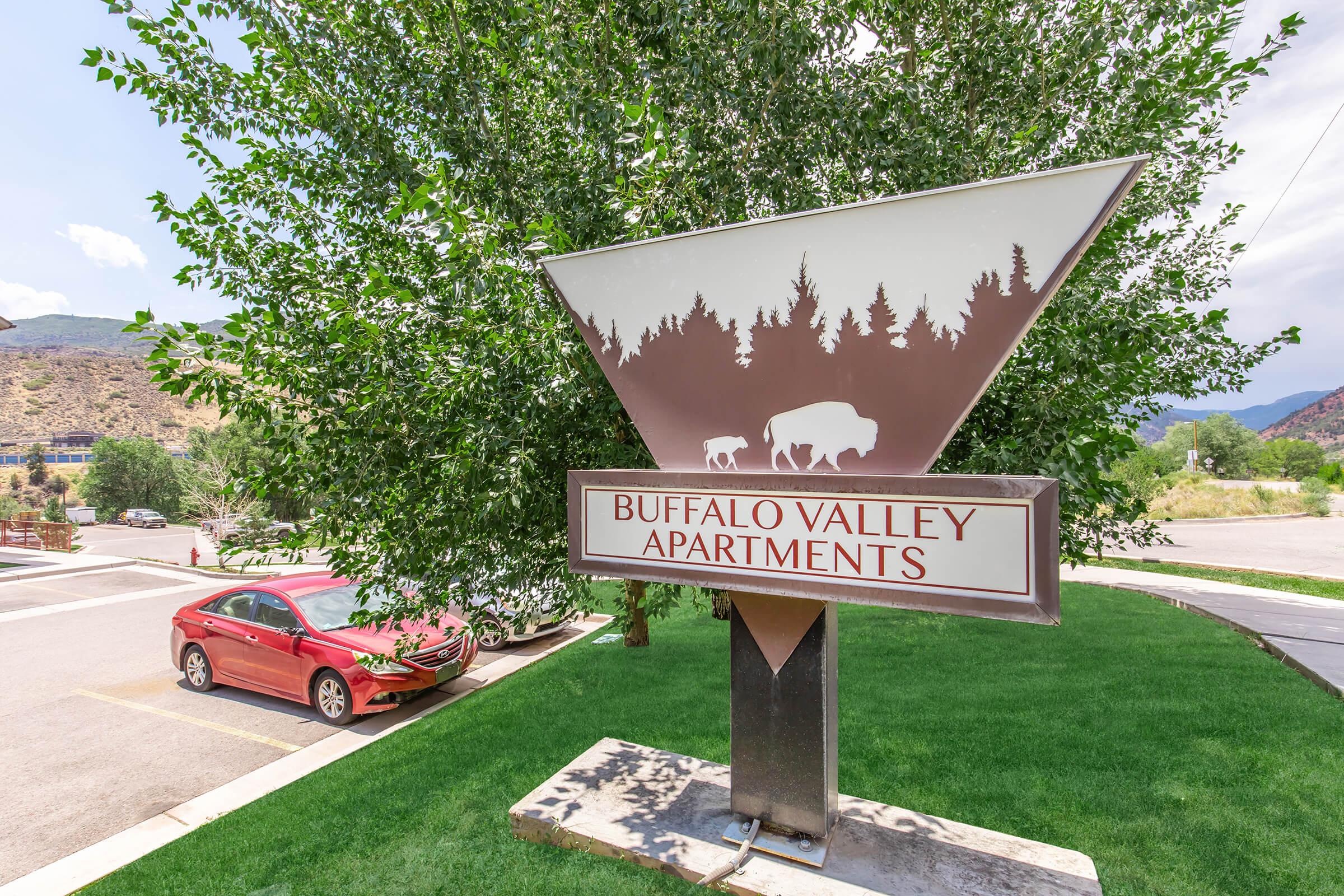 Sign for Buffalo Valley Apartments displayed on a triangular monument. The sign features a silhouette of a buffalo and is surrounded by greenery, with a car parked nearby. The background shows a mountainous landscape under a partly cloudy sky.