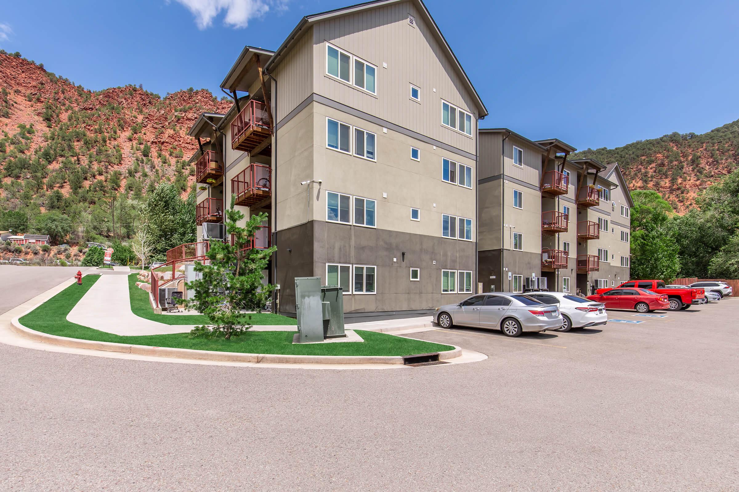 A modern multi-story residential building situated in a scenic area with rocky red hills in the background. The structure features balconies and a landscaped walkway. Several cars are parked along a paved lot, with greenery around the edges, under a clear blue sky.