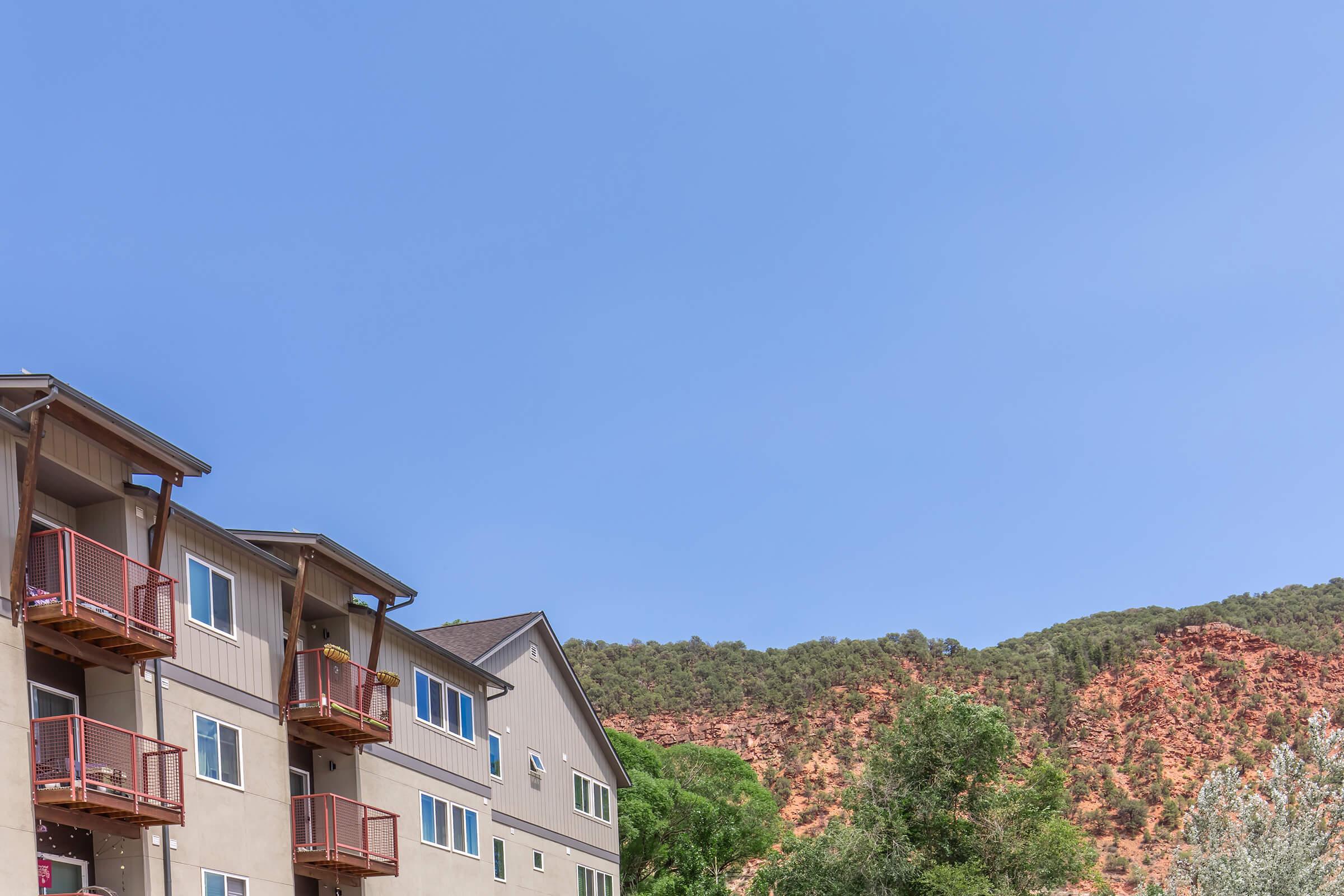 A multi-story apartment building with balconies, situated in a natural setting. In the background, there are red rock formations and green trees under a clear blue sky.