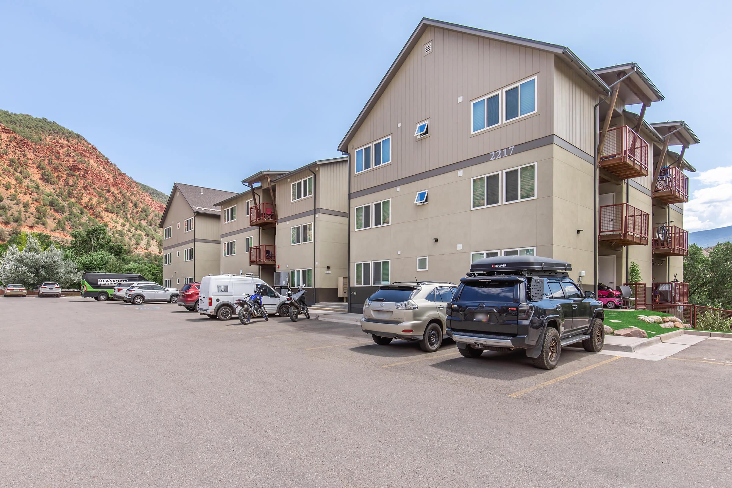 A modern multi-story apartment building with multiple balconies, surrounded by mountains. The parking lot features several vehicles, including a motorcycle and a van. Lush greenery is visible nearby, indicating a natural setting. The sky is clear with a few clouds.