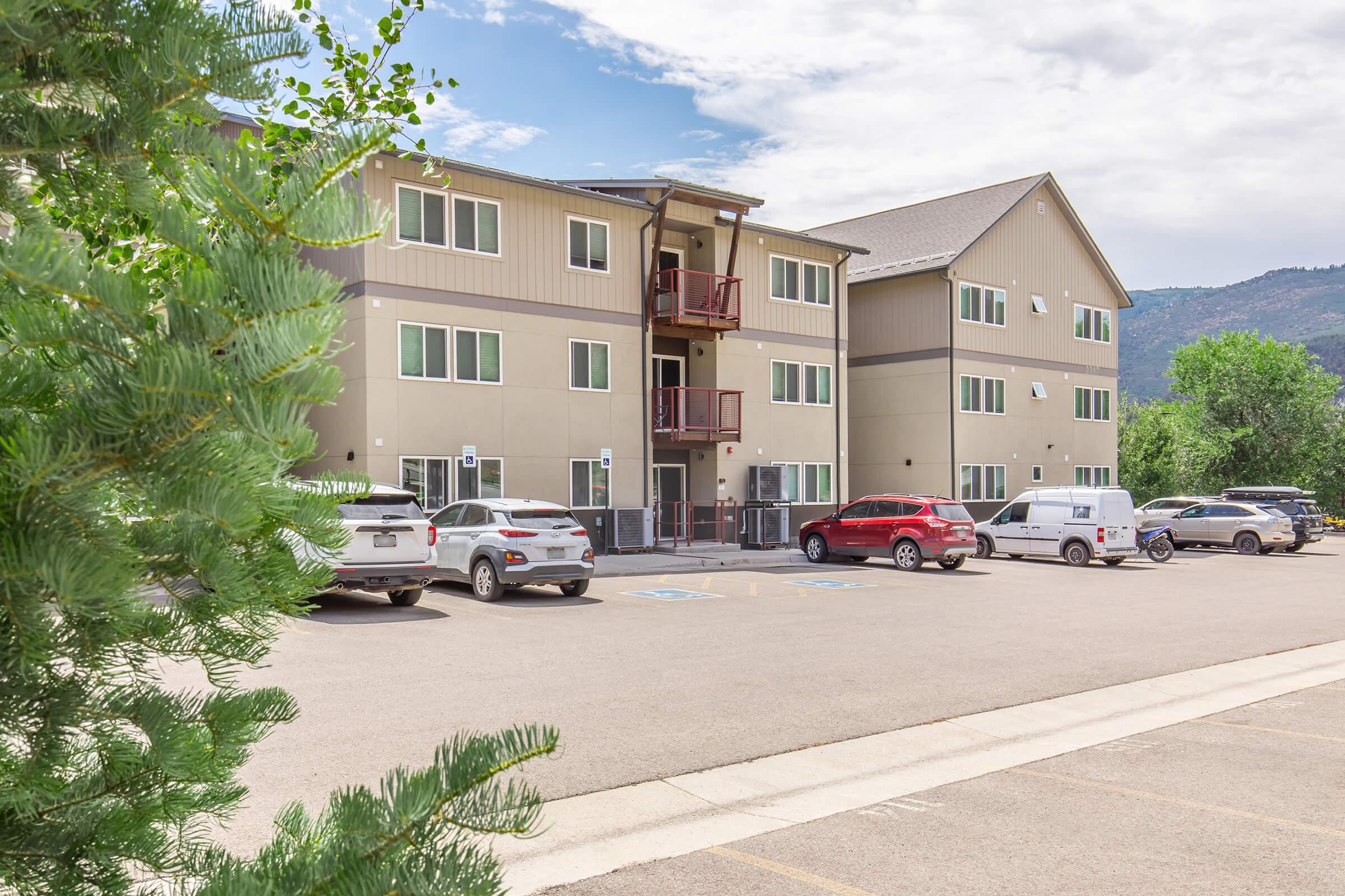 A three-story residential building with modern architecture, featuring balconies and parked cars in a lot. In the background, there are trees and mountains under a partly cloudy sky. The setting appears to be a suburban area with ample parking space.