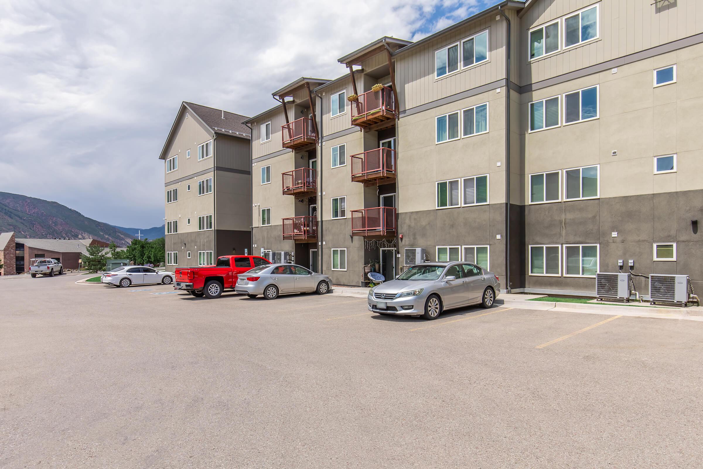 Modern apartment building with three stories, featuring multiple balconies. In the foreground, there are several parked cars including a red pickup truck and a silver sedan. The background shows a mountainous landscape under a partly cloudy sky.