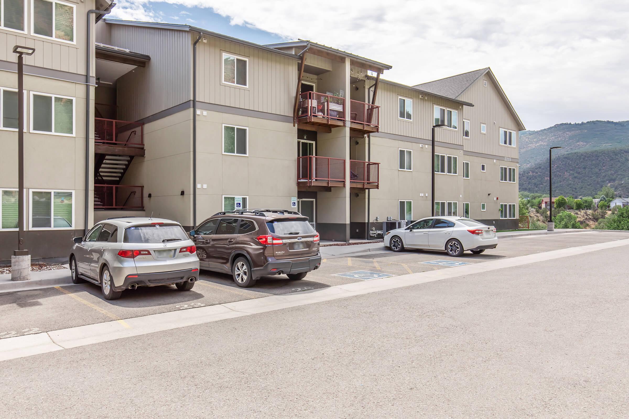 A modern multi-story apartment building with balconies, surrounded by parking spaces. Several vehicles are parked in front, and mountains are visible in the background under a partly cloudy sky.