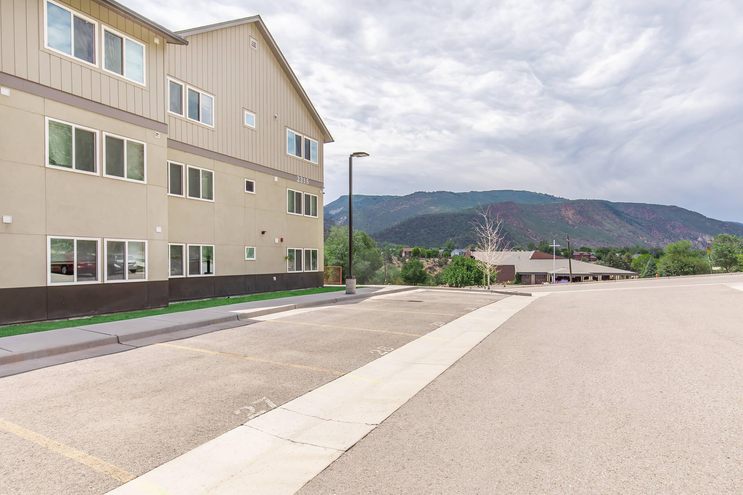 A view of a modern building with large windows, situated near a parking area and surrounded by green hills. The sky is partly cloudy, and there are residential houses visible in the background. The overall scene depicts a suburban landscape with a focus on the building's architecture.