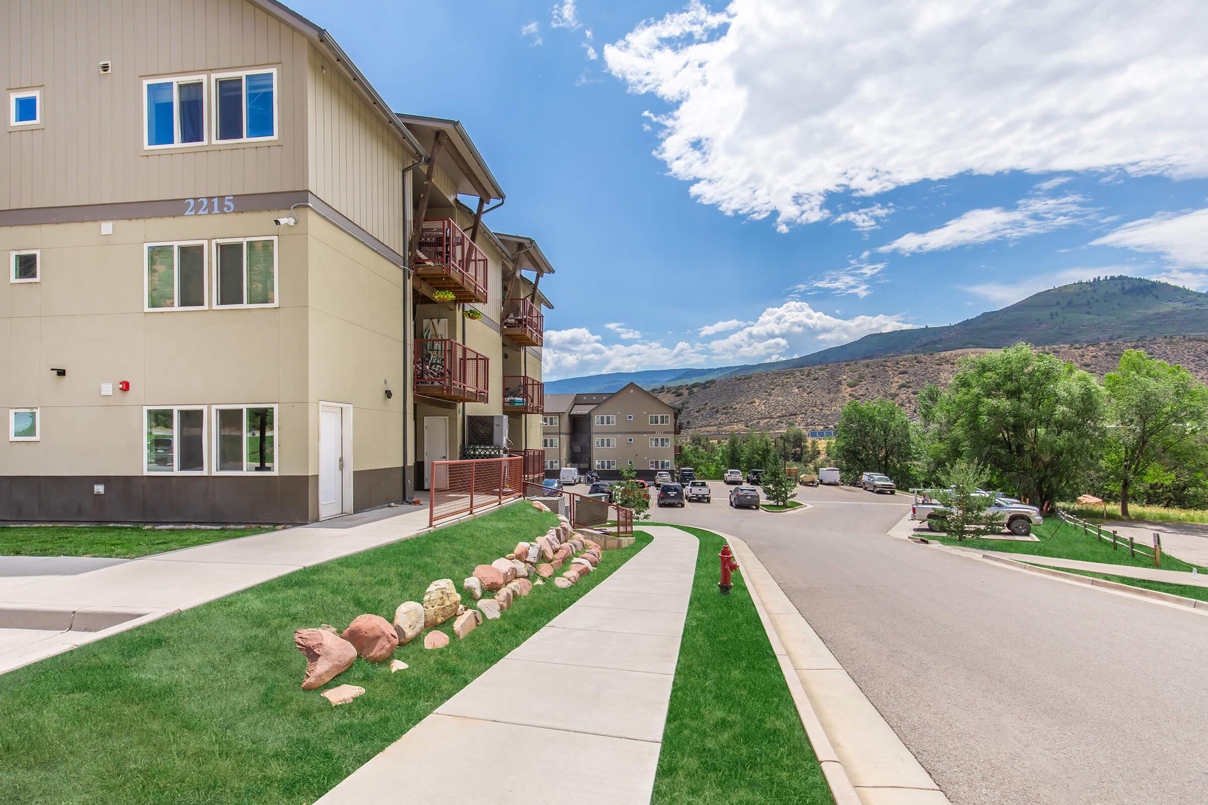 A residential area featuring modern multi-family buildings. The foreground includes a landscaped pathway bordered by rocks, while the background showcases a scenic view of mountains under a partly cloudy sky. Vehicles are parked along the street, and green trees add to the neighborhood's ambiance.