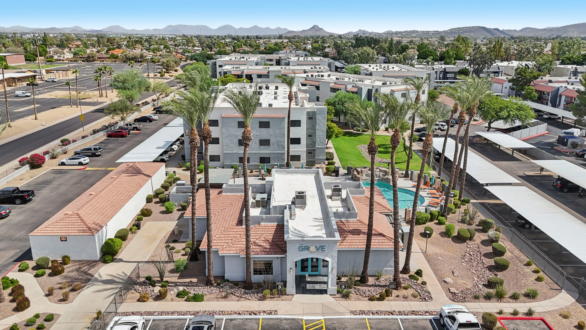 Aerial view of a residential complex featuring several buildings, palm trees, and a swimming pool. The entrance is prominently labeled "GROVE," with surrounding parking areas and landscaped grounds. In the background, mountains and additional urban structures are visible, indicating a suburban setting.
