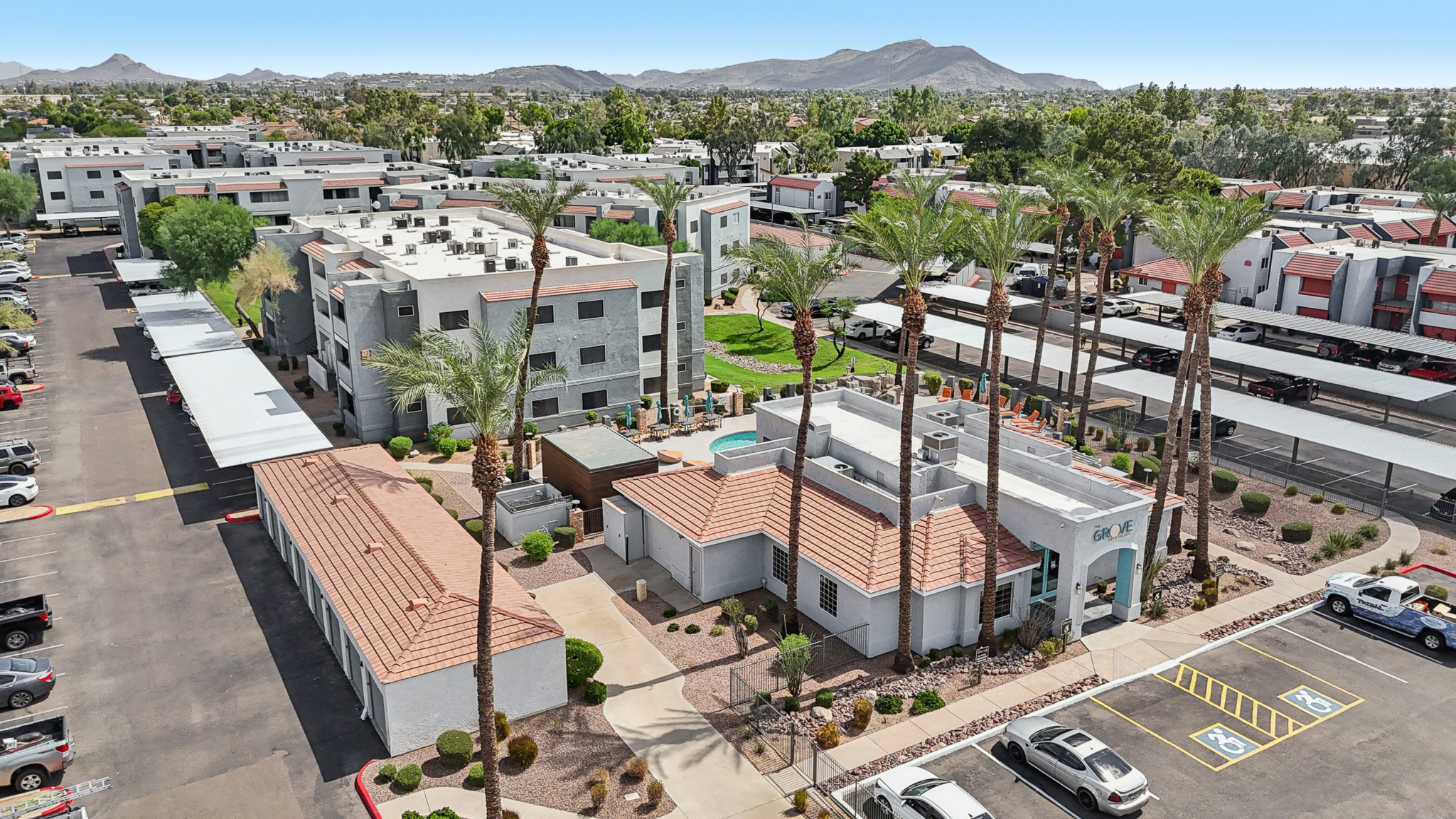 Aerial view of a residential community featuring several buildings with terracotta roofs, palm trees, and a landscaped area. Parking spaces are visible, along with a swimming pool area. In the background, additional residential structures and mountains can be seen under a clear blue sky.