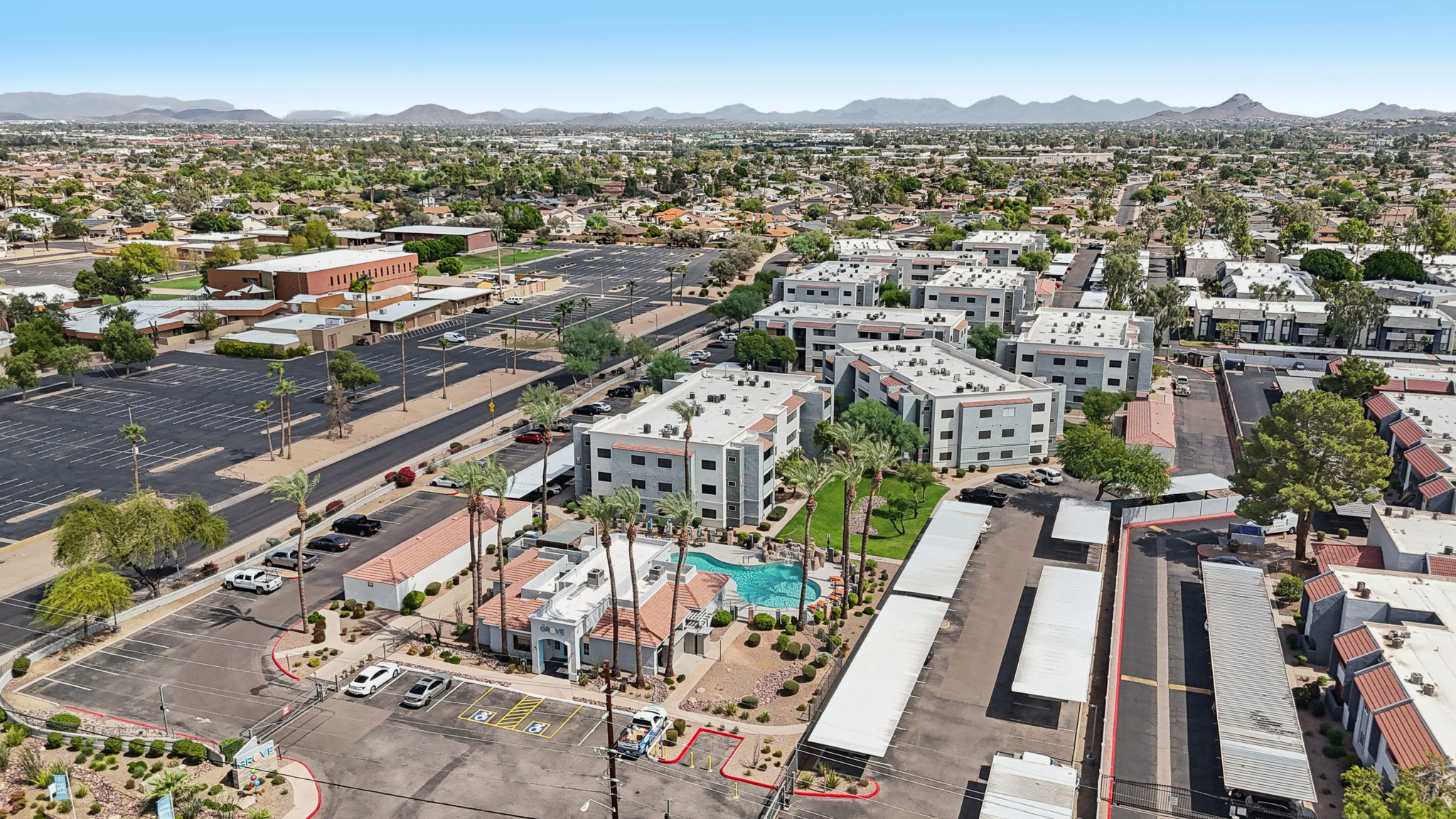 Aerial view of a suburban area featuring residential buildings, a swimming pool, and parking lots. Surrounding mountains can be seen in the background, along with a clear blue sky. The landscape includes palm trees and a mix of commercial and residential structures.