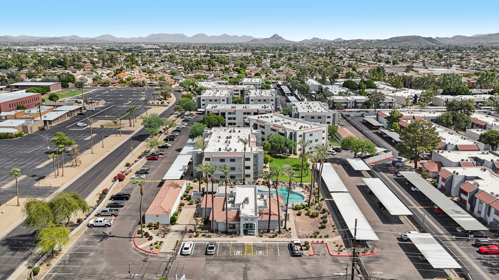 Aerial view of a residential area featuring multiple low-rise buildings with a central green space and swimming pool. Surrounding the complex are parking areas and a mix of other structures, with mountains visible in the distance against a clear blue sky.