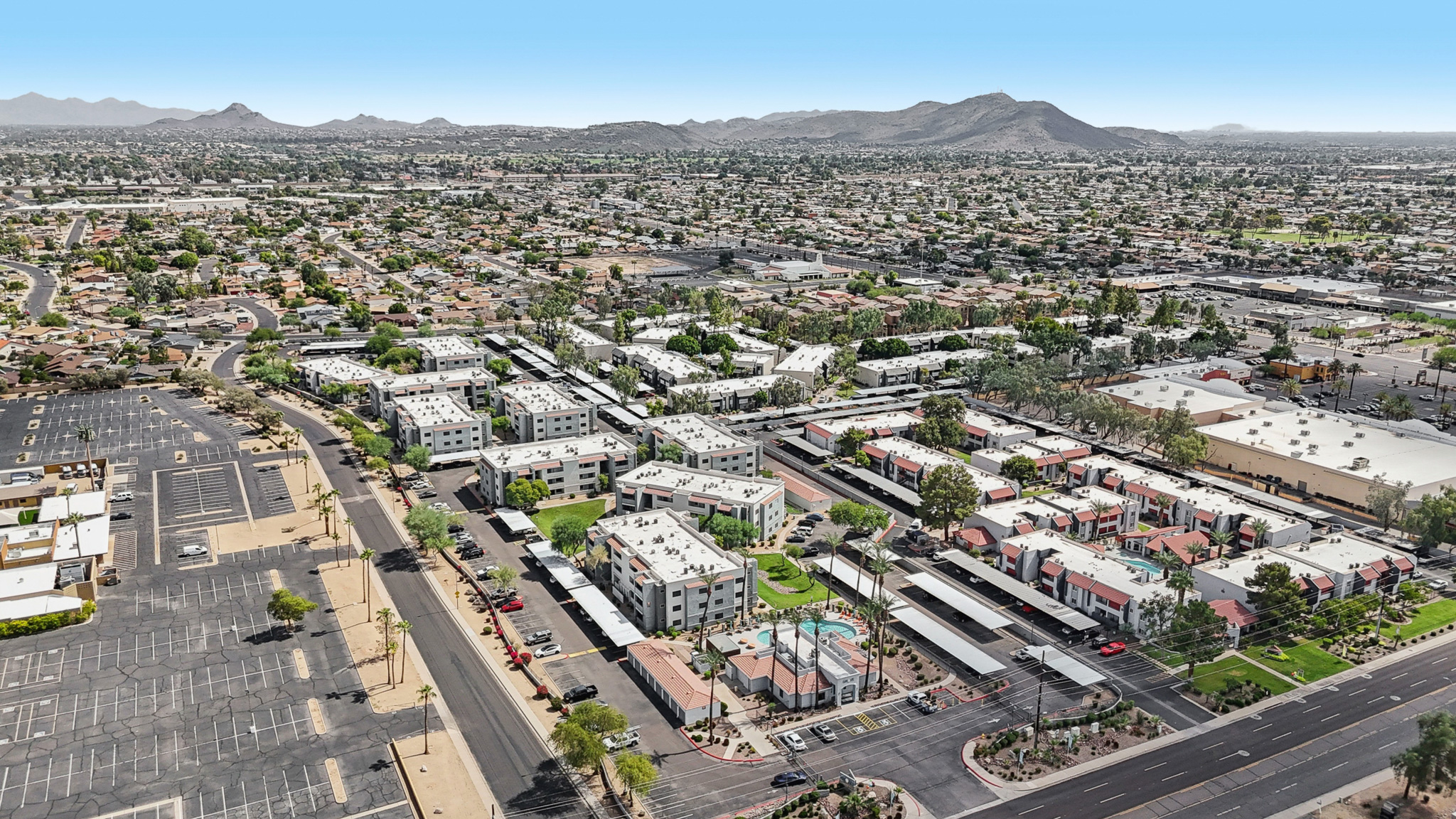 Aerial view of a suburban area featuring multiple residential buildings with swimming pools, surrounded by roads and parking lots. In the background, mountains are visible under a clear blue sky, highlighting the expansive layout of the neighborhood and its proximity to urban amenities.