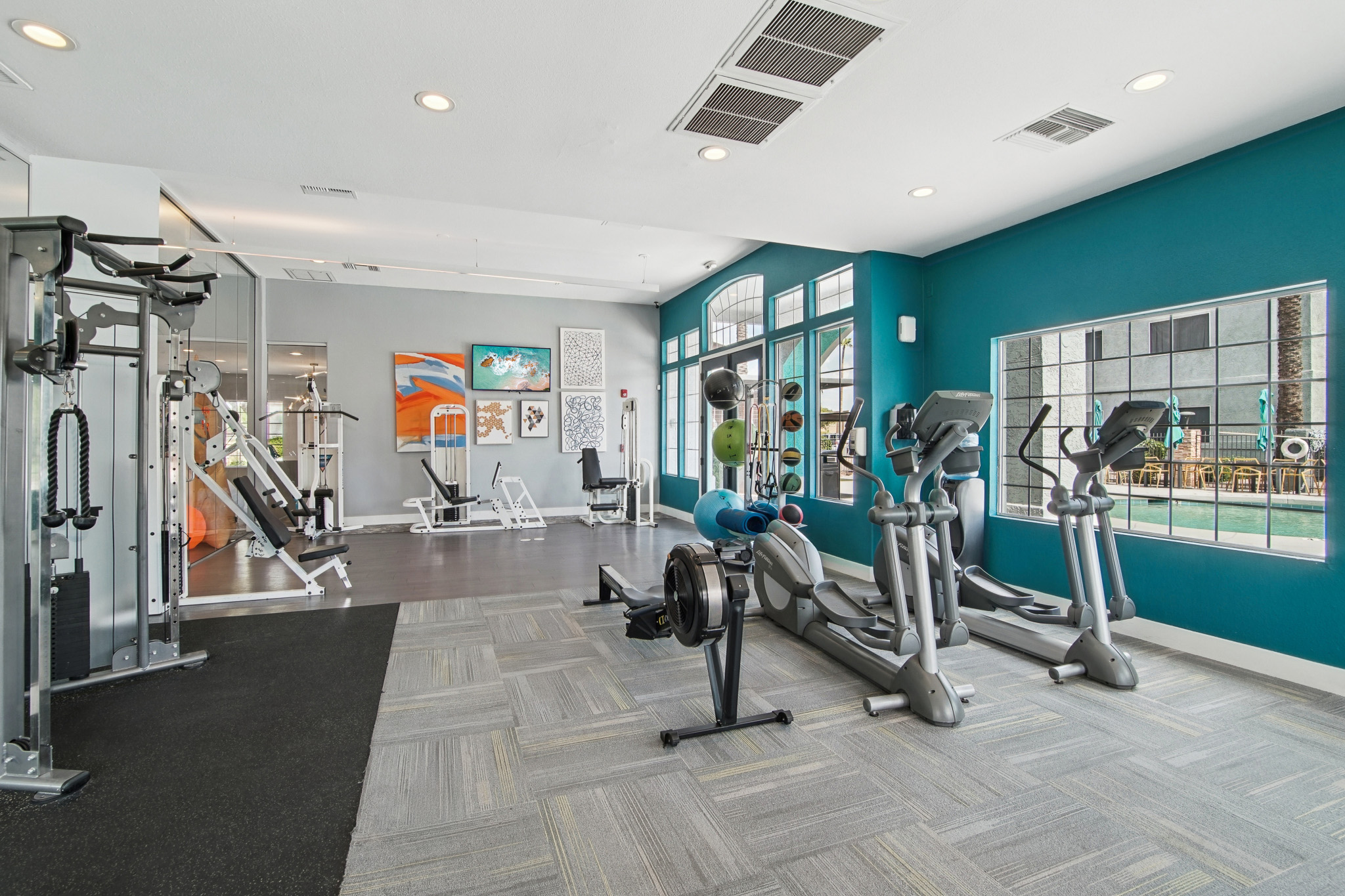 A modern gym interior featuring various exercise equipment, including treadmills, elliptical machines, and weight machines. The space is brightly lit with large windows, and colorful artwork is displayed on the walls. The flooring is a mix of carpet and tile, creating a welcoming fitness environment.