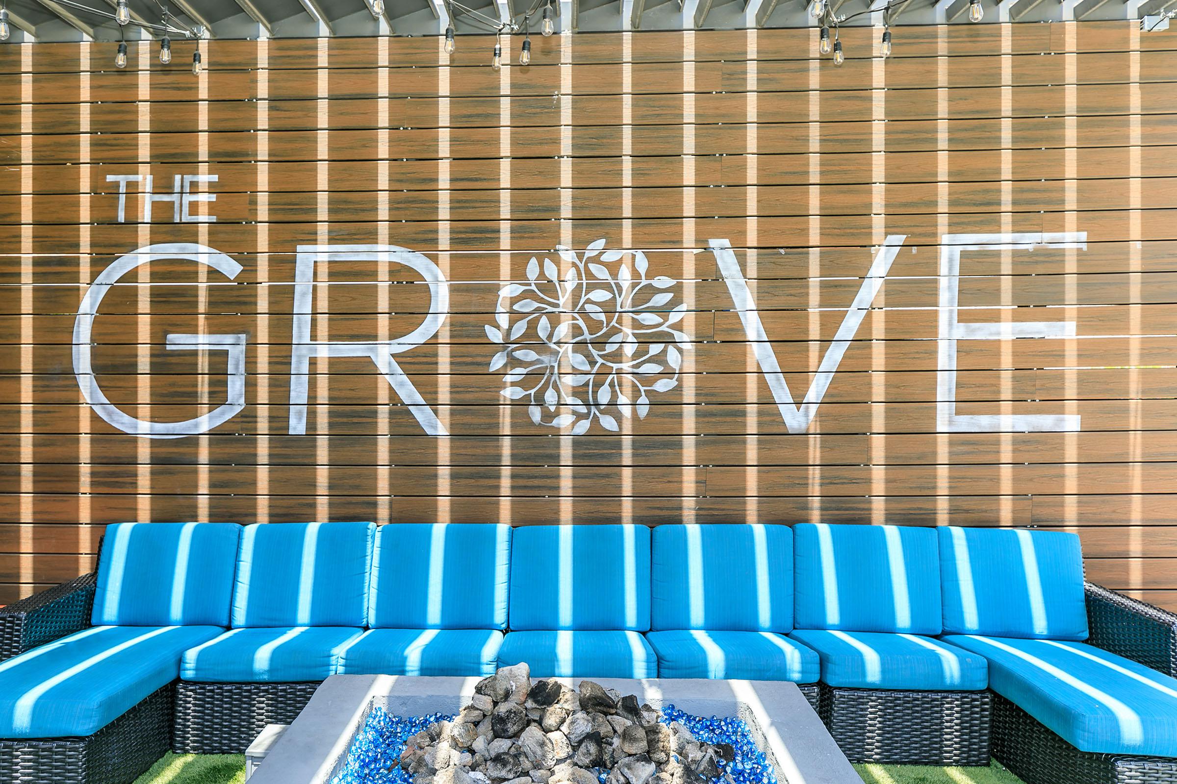 A cozy outdoor seating area featuring a bright blue couch in front of a wooden wall. The wall has the text "THE GROVE" prominently displayed, accompanied by a decorative design of a flower or tree. A fire pit with stones is positioned in front of the seating area, enhancing the inviting ambiance.