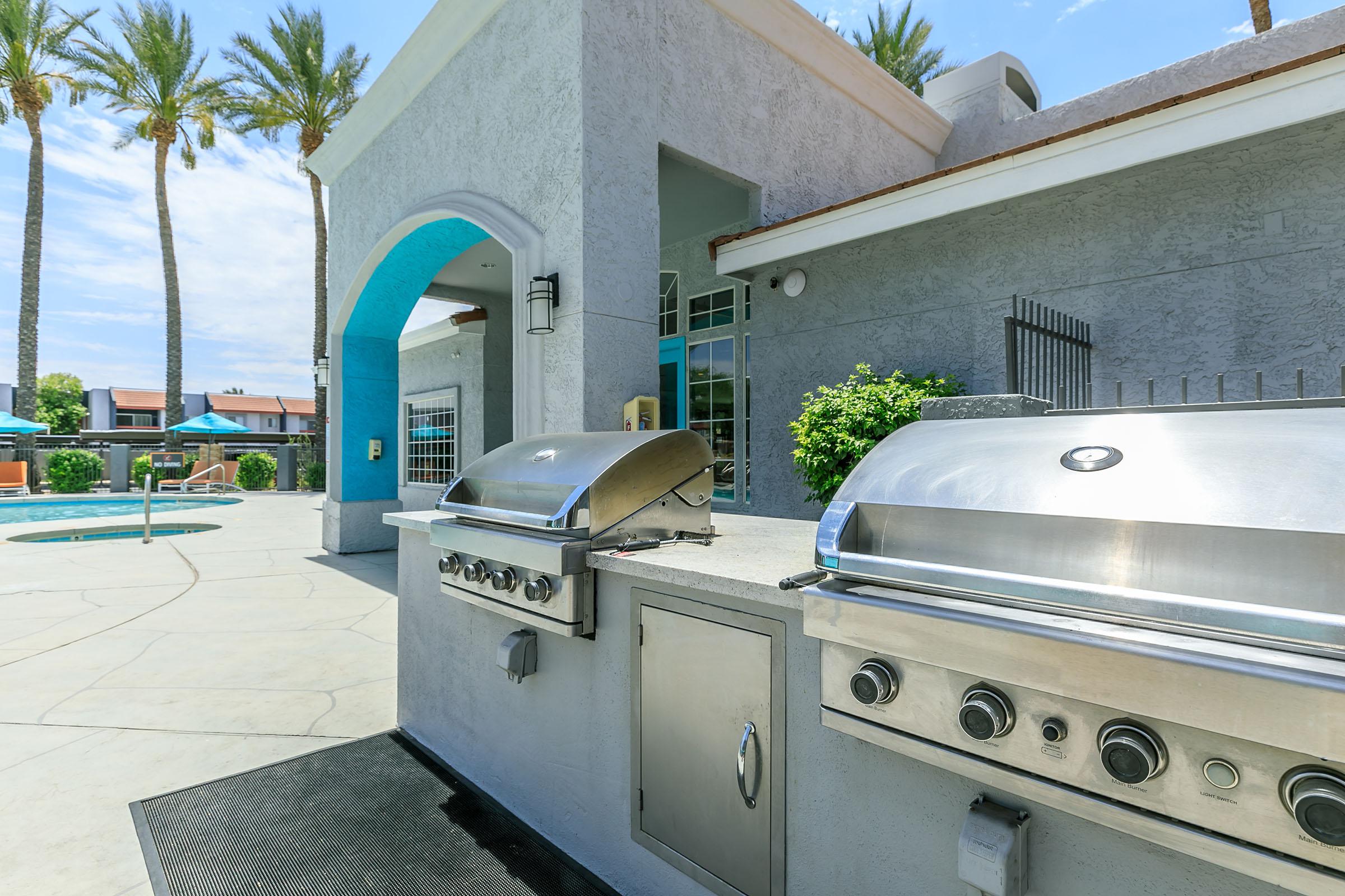 A bright outdoor area featuring two stainless steel gas grills beside a pool. The scene includes palm trees in the background and a blue archway. The griddles are set up for barbecuing, with a modern architectural design of the surrounding building. Ideal for gatherings and outdoor cooking.