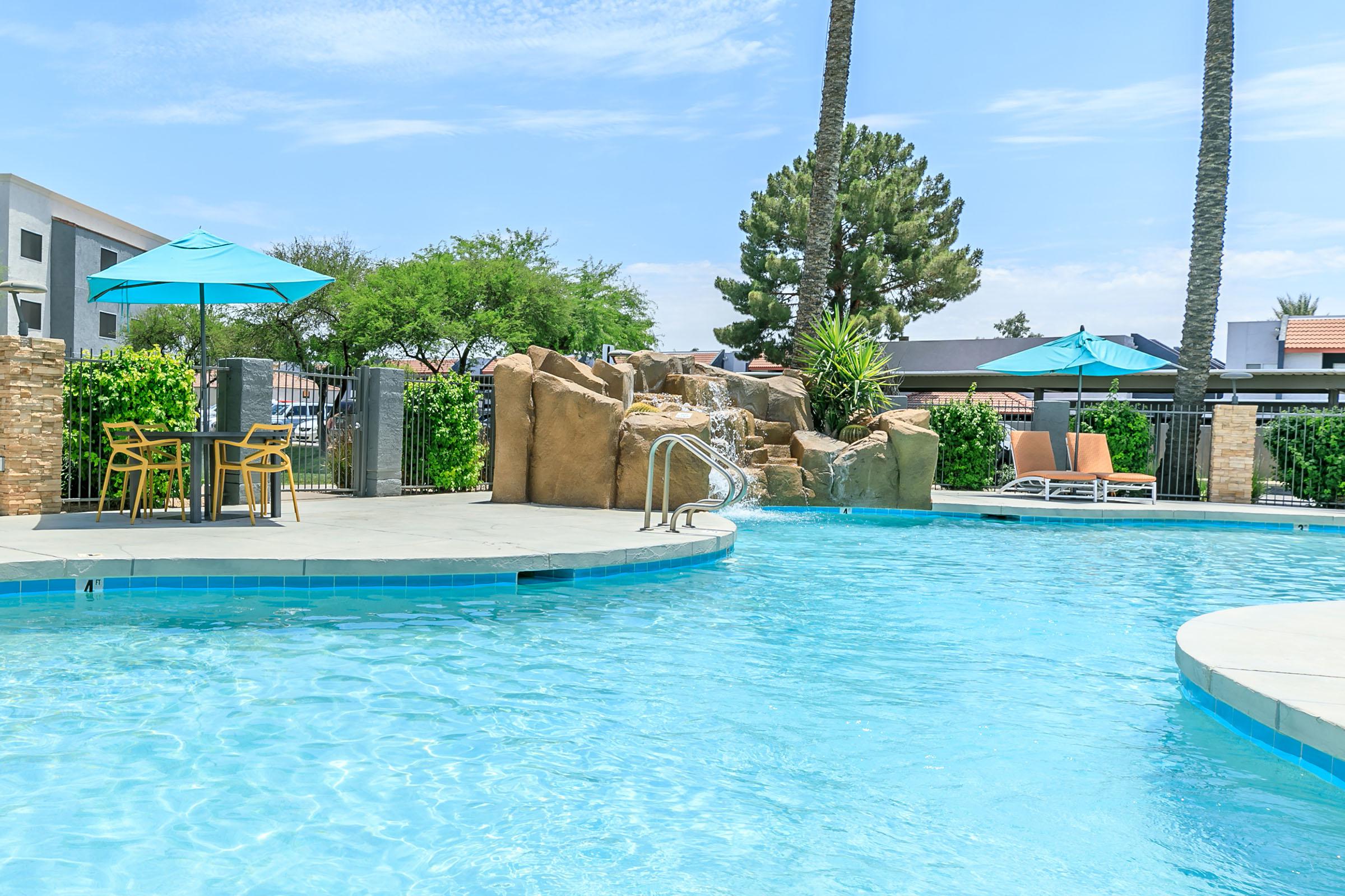 A sunny outdoor pool area featuring clear blue water, a rock formation waterfall, and several palm trees. There are yellow lounge chairs by the pool, turquoise umbrellas over tables, and lush greenery surrounding the space, creating a relaxing and inviting atmosphere.