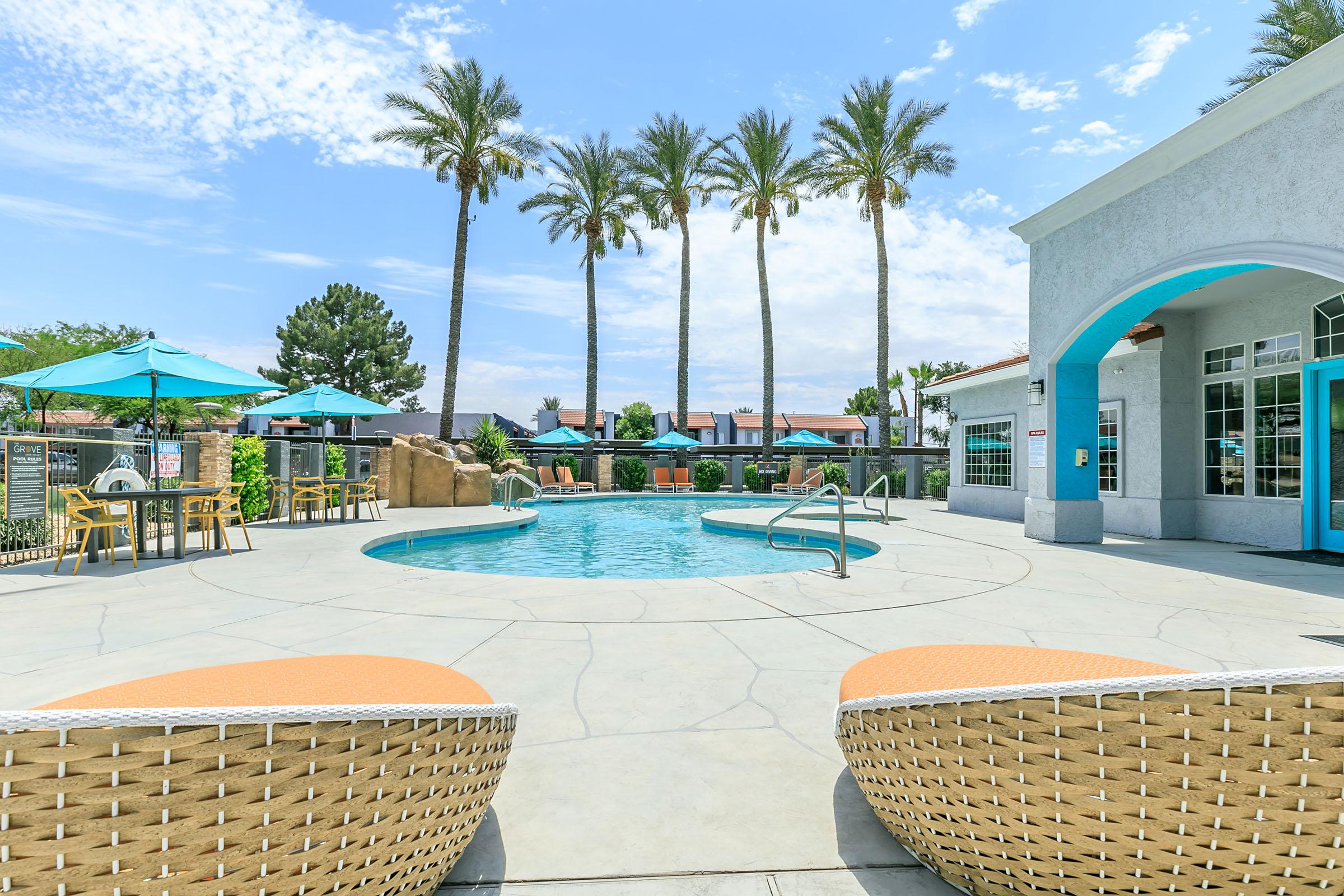 A sunny pool area featuring a clear blue swimming pool surrounded by palm trees. There are two stylish lounge chairs in the foreground, and vibrant umbrellas over seating areas. The backdrop includes a light-colored building with large windows, creating a relaxed outdoor atmosphere.