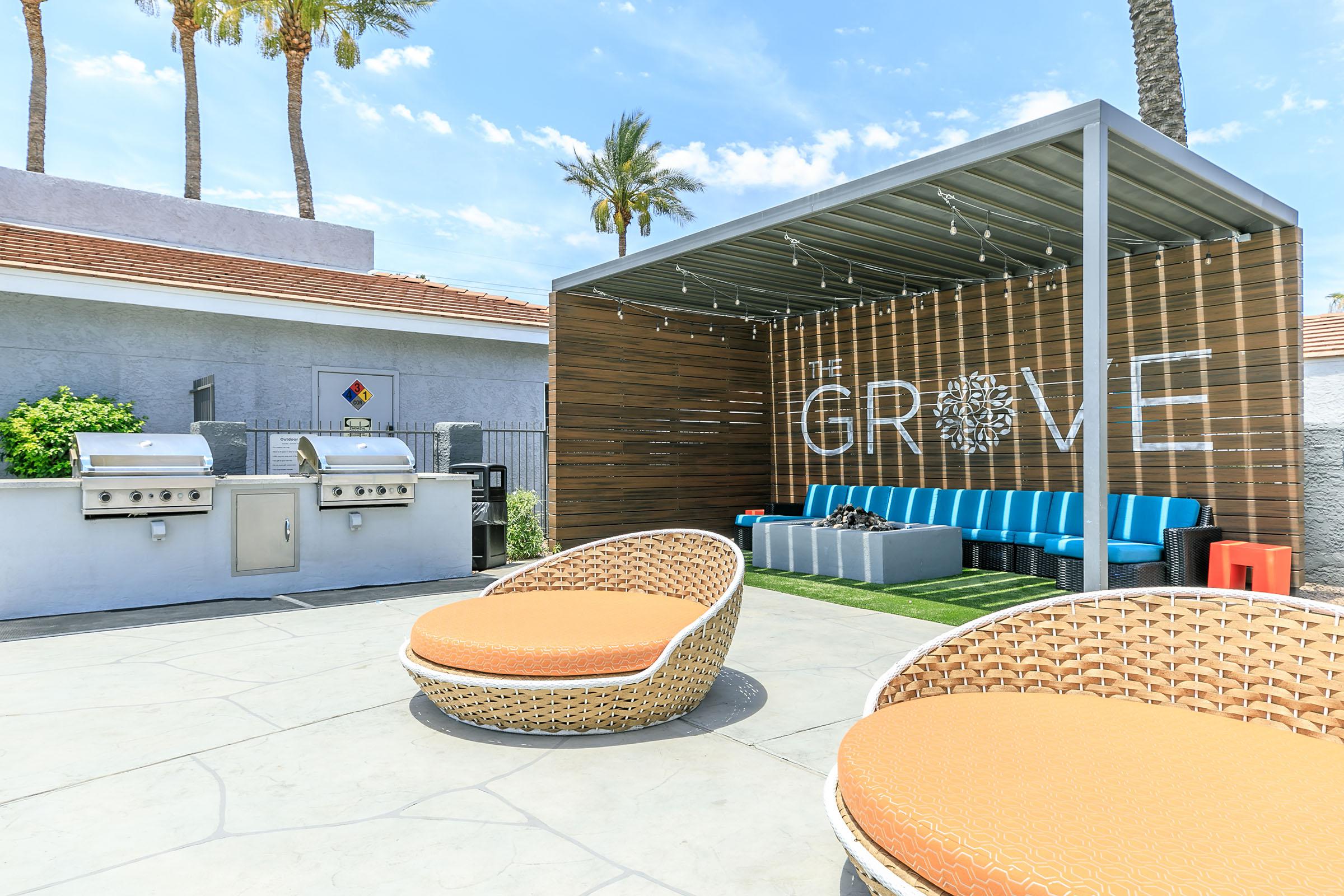 A sunny outdoor area featuring a shaded seating space with a large sign reading "THE GROVE." There are comfortable blue couches, stylish circular chairs with orange cushions, and a barbecue grill setup nearby, surrounded by palm trees under a clear blue sky.