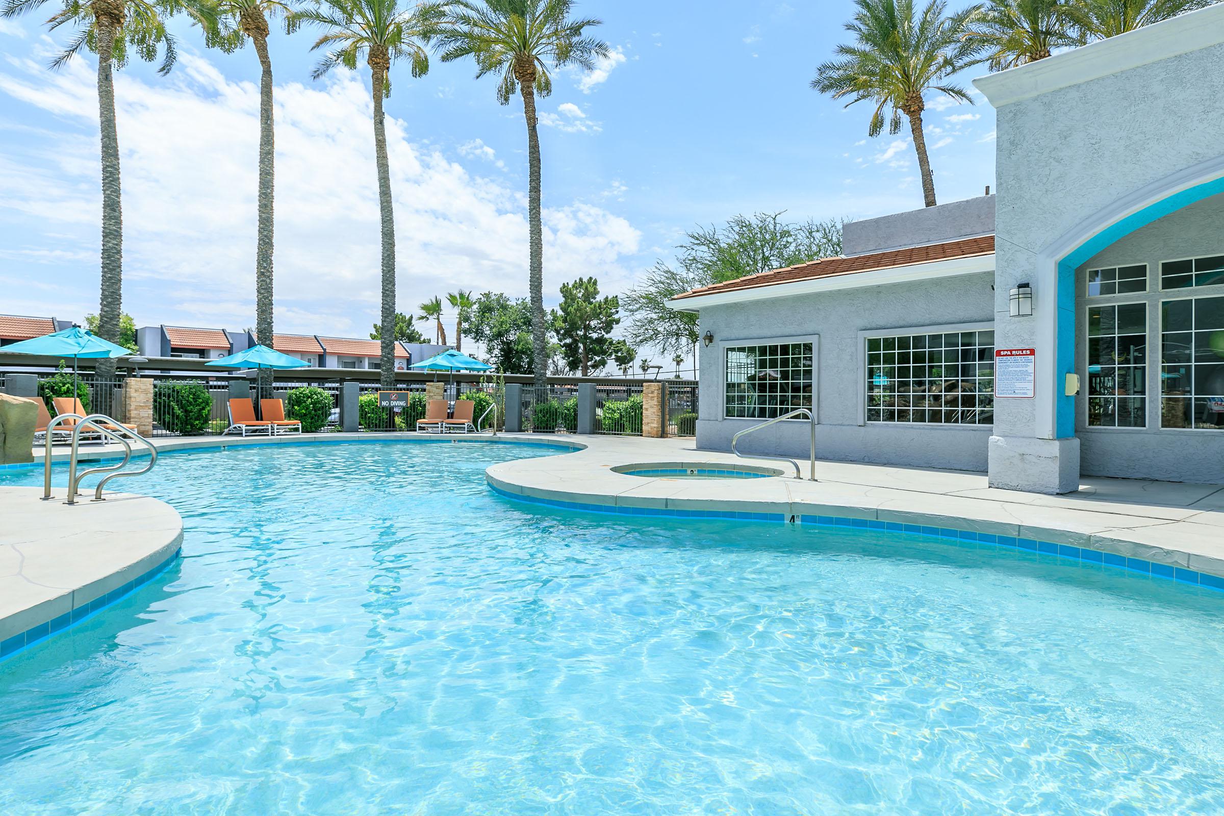 A clear blue swimming pool surrounded by palm trees and lounge chairs. One section of the pool features a built-in hot tub. In the background, there is a building with large windows and outdoor seating areas, with umbrellas providing shade. The sky is bright and partly cloudy.