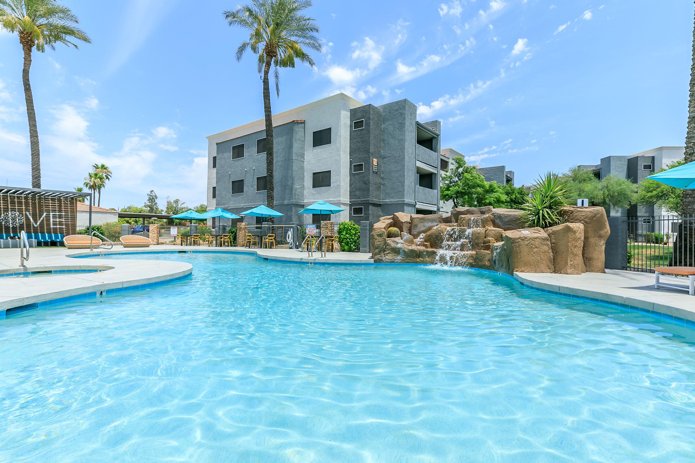 A bright outdoor swimming pool with clear blue water, surrounded by palm trees and lounge chairs. In the background, a modern apartment building with gray and white facades. A small rock waterfall adds a tropical touch, and several teal umbrellas provide shade for guests enjoying the space.