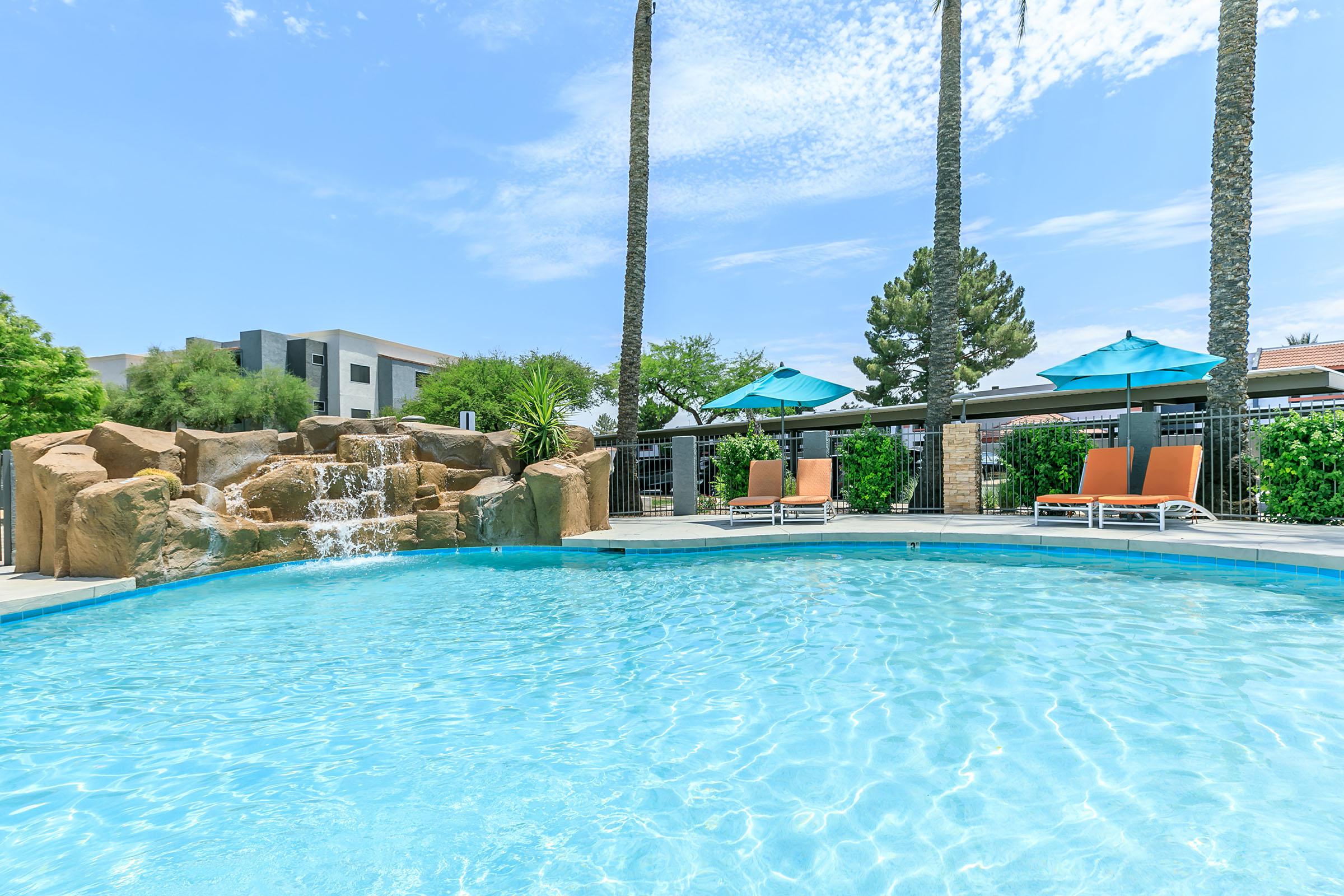 A serene pool surrounded by palm trees, featuring a rocky waterfall on one side. Two turquoise umbrellas shade orange lounge chairs near the water's edge. In the background, modern buildings and greenery can be seen under a bright blue sky with scattered clouds.