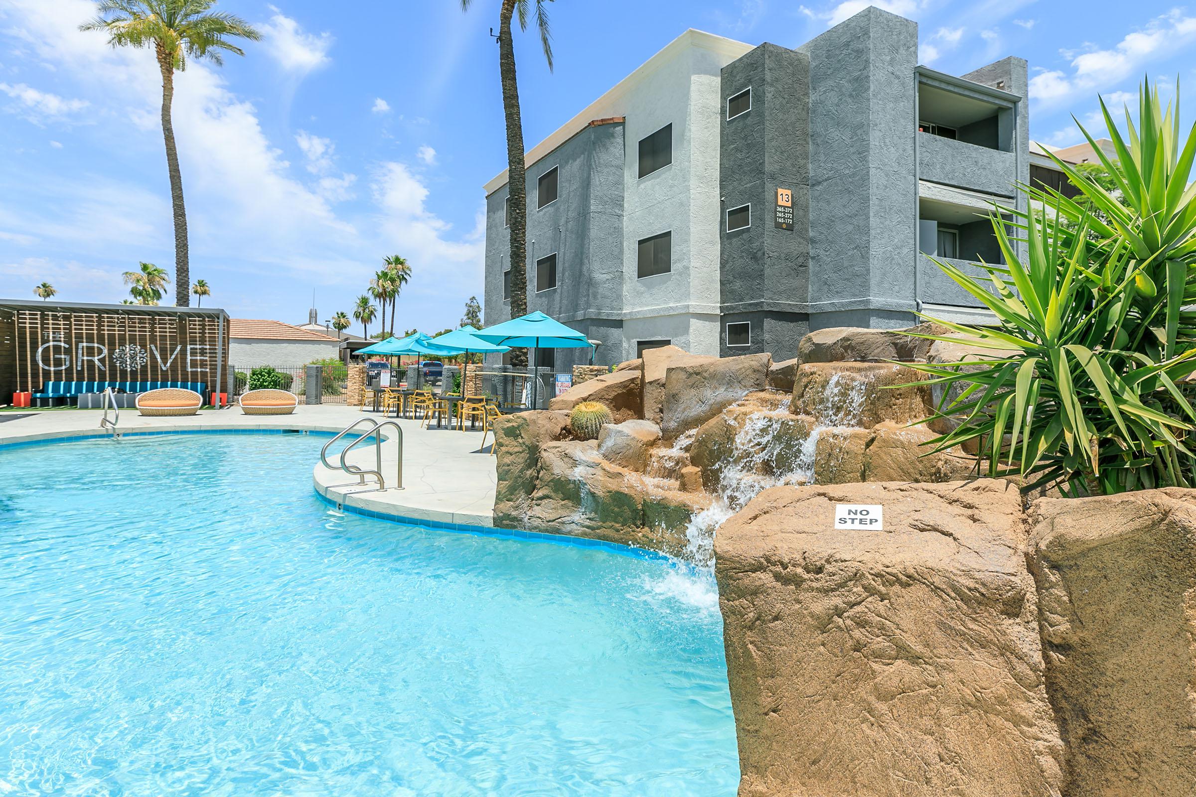 A sunny outdoor pool area featuring a rock waterfall, surrounded by palm trees and chaise lounges. The pool has a shallow entry and is marked with a "No Step" sign. In the background, there is a multi-story building with balconies and several blue umbrellas for shade.